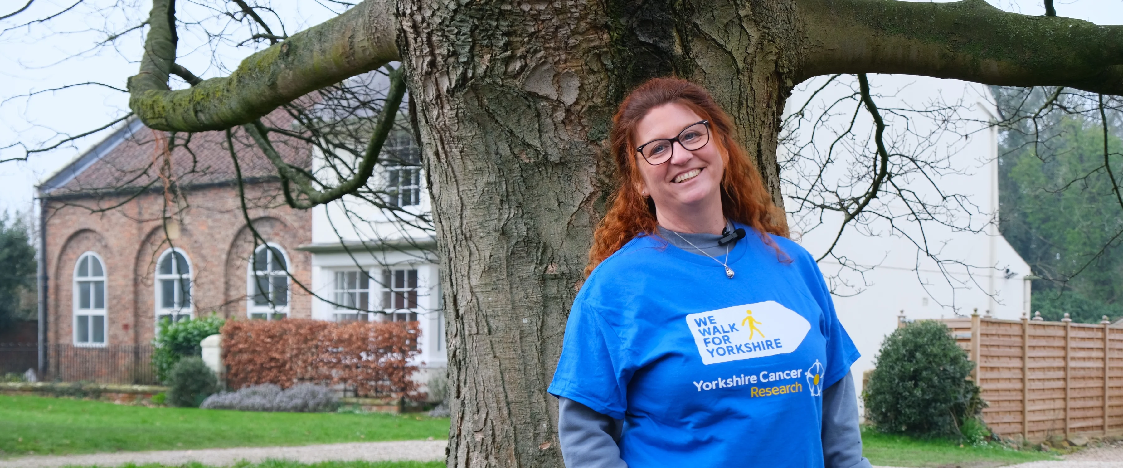 Jackie is smiling at the camera. She has red hair and is wearing black glasses. Behind her is a large tree and some daffodils.