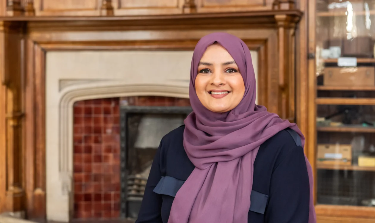 Saima is wearing a purple hijab and standing in front of an antique wooden fireplace.