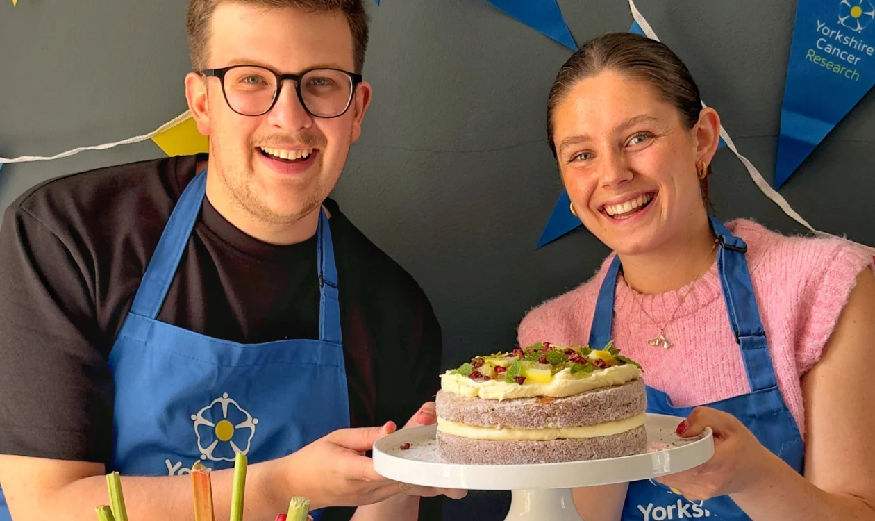 Two people holding a rhubarb doughnut cake. They are wearing blue aprons and behind them is bright charity-branded bunting.
