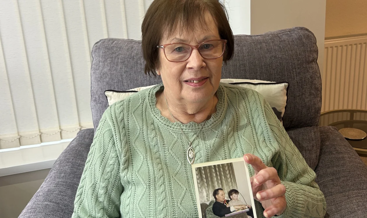 A lady holding a family photograph. She is wearing a green jumper and sitting on a grey sofa.