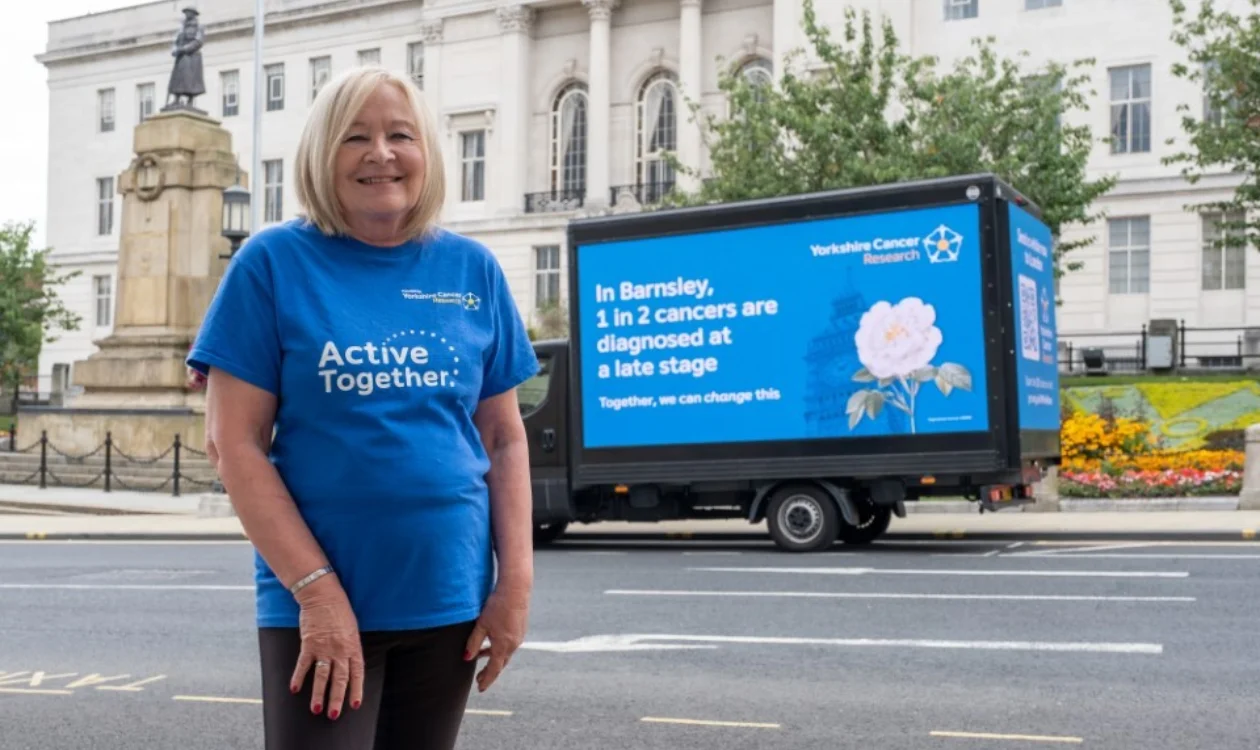 A lady wearing a blue "Active Together" t-shirt standing outside a large white building in Barnsley. behind her is a digital van that says "In Barnsley 1 in 2 cancers are diagnosed at a late stage"