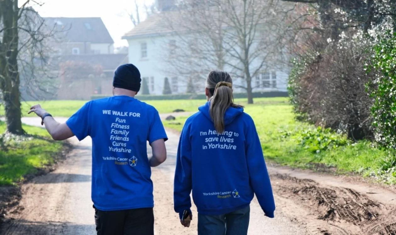 Two people wearing blue branded We Walk for Yorkshire tops are walking outside on a crisp spring day. They are facing away from the camera and in front of them is a large white building and trees.