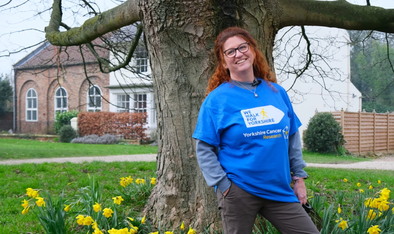 Jackie is smiling at the camera. She has red hair and is wearing black glasses. Behind her is a large tree and some daffodils.