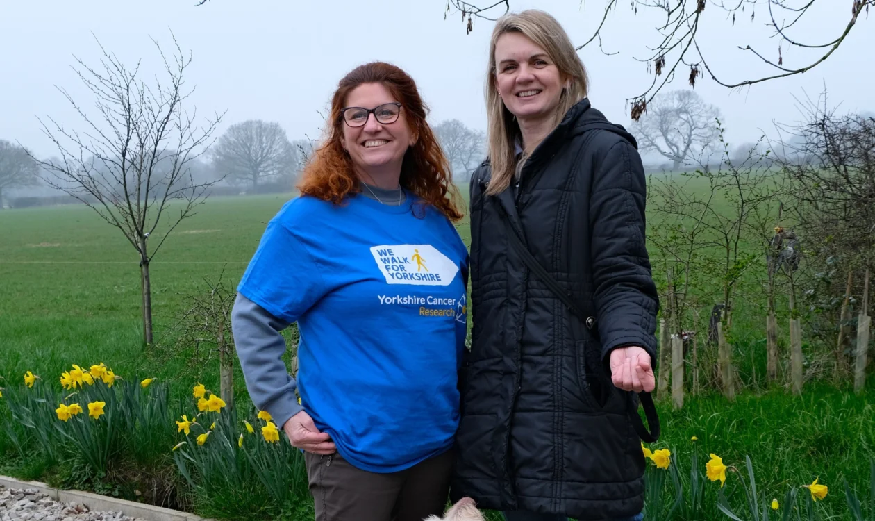 Jackie and her friend are stood in front of a large grassy field. Behind them are some daffodils. Jackie is wearing a blue 'We Walk for Yorkshire' T-shirt.