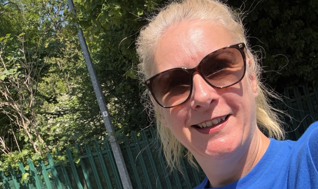 Jayne in a selfie. She is stood outside on a sunny day. She is wearing black sunglasses.