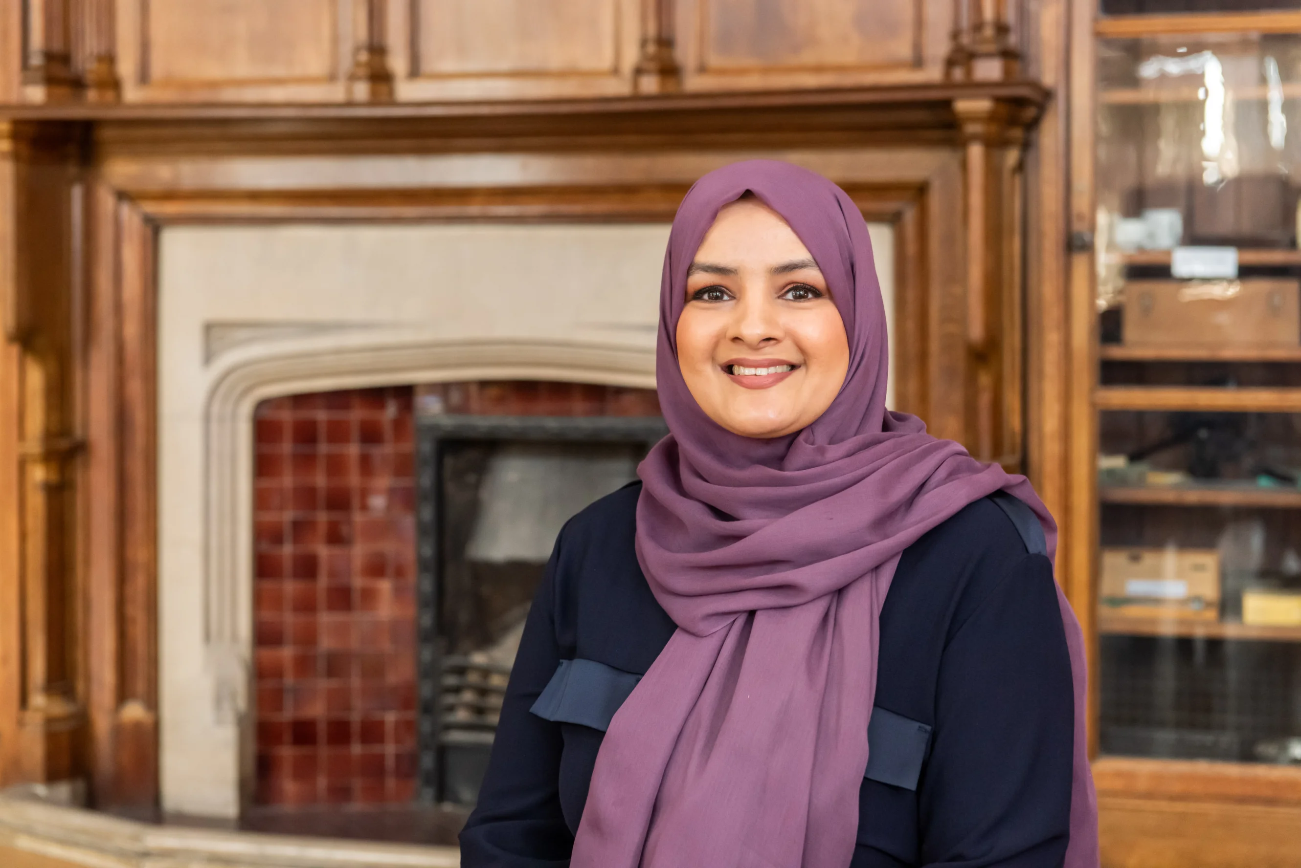 Saima is wearing a purple hijab and standing in front of an antique wooden fireplace.
