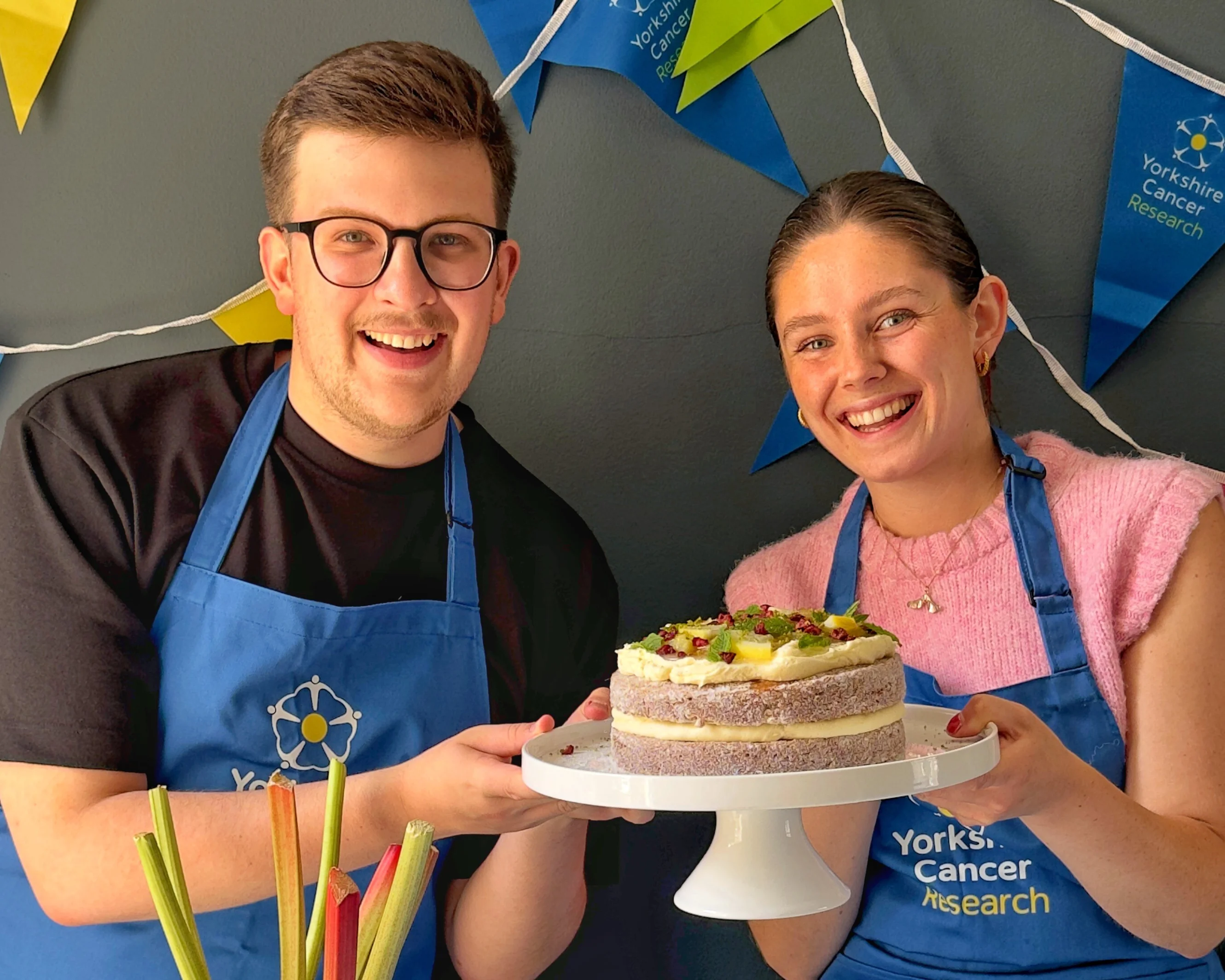 Two people holding a rhubarb doughnut cake. They are wearing blue aprons and behind them is bright charity-branded bunting.