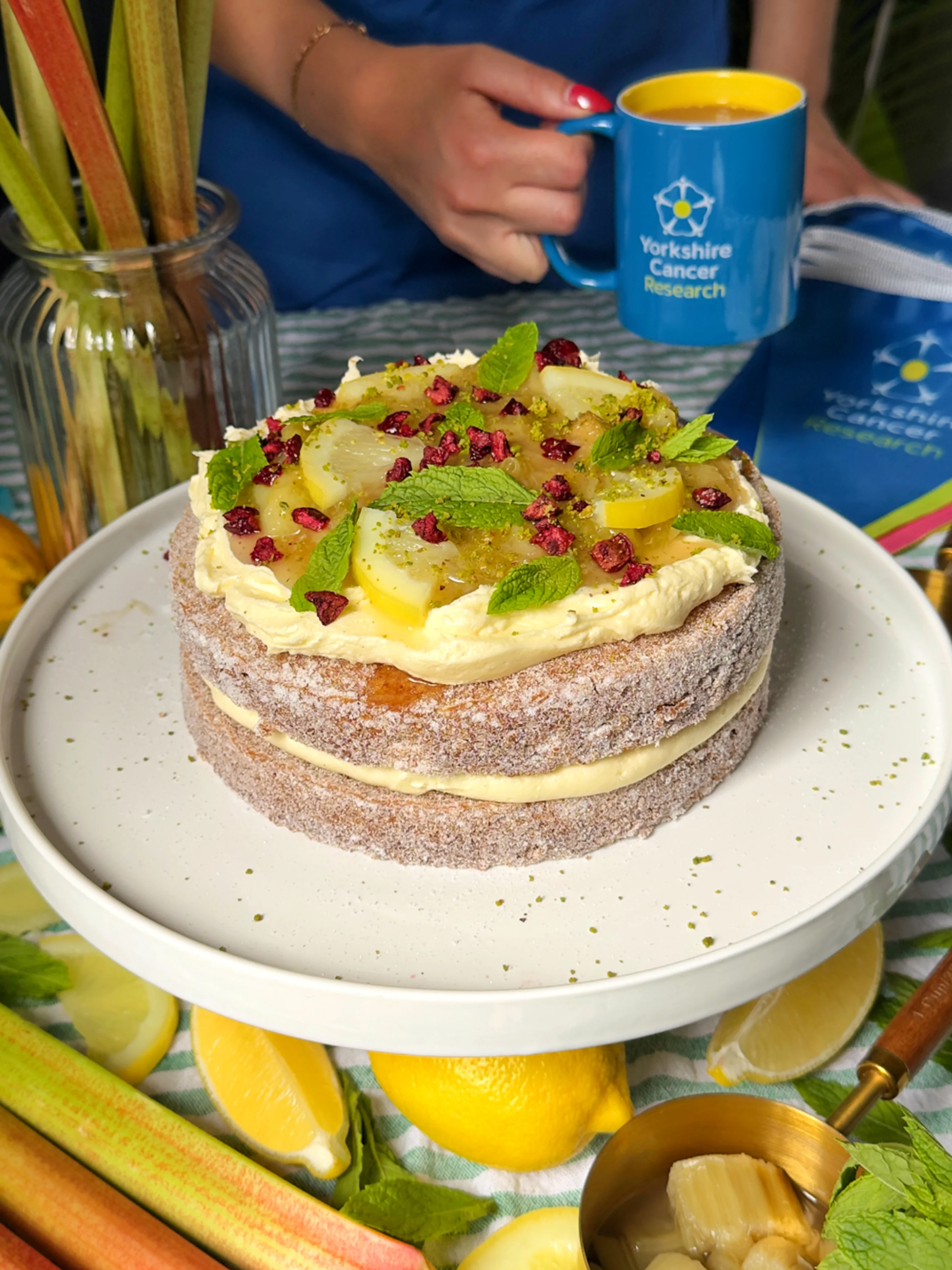 An image of a rhubarb and lemon doughnut cake decorated with slices of lemon and pomegranate seeds. In the background is a blue Yorkshire Cancer Research mug.