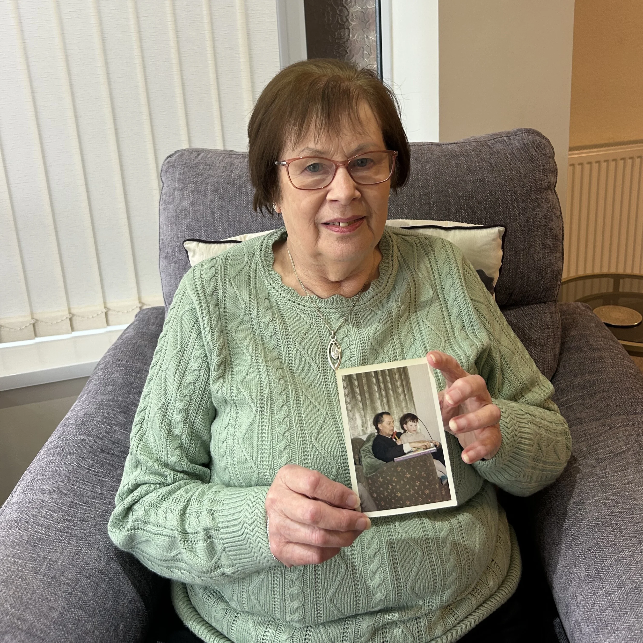 A lady holding a family photograph. She is wearing a green jumper and sitting on a grey sofa.