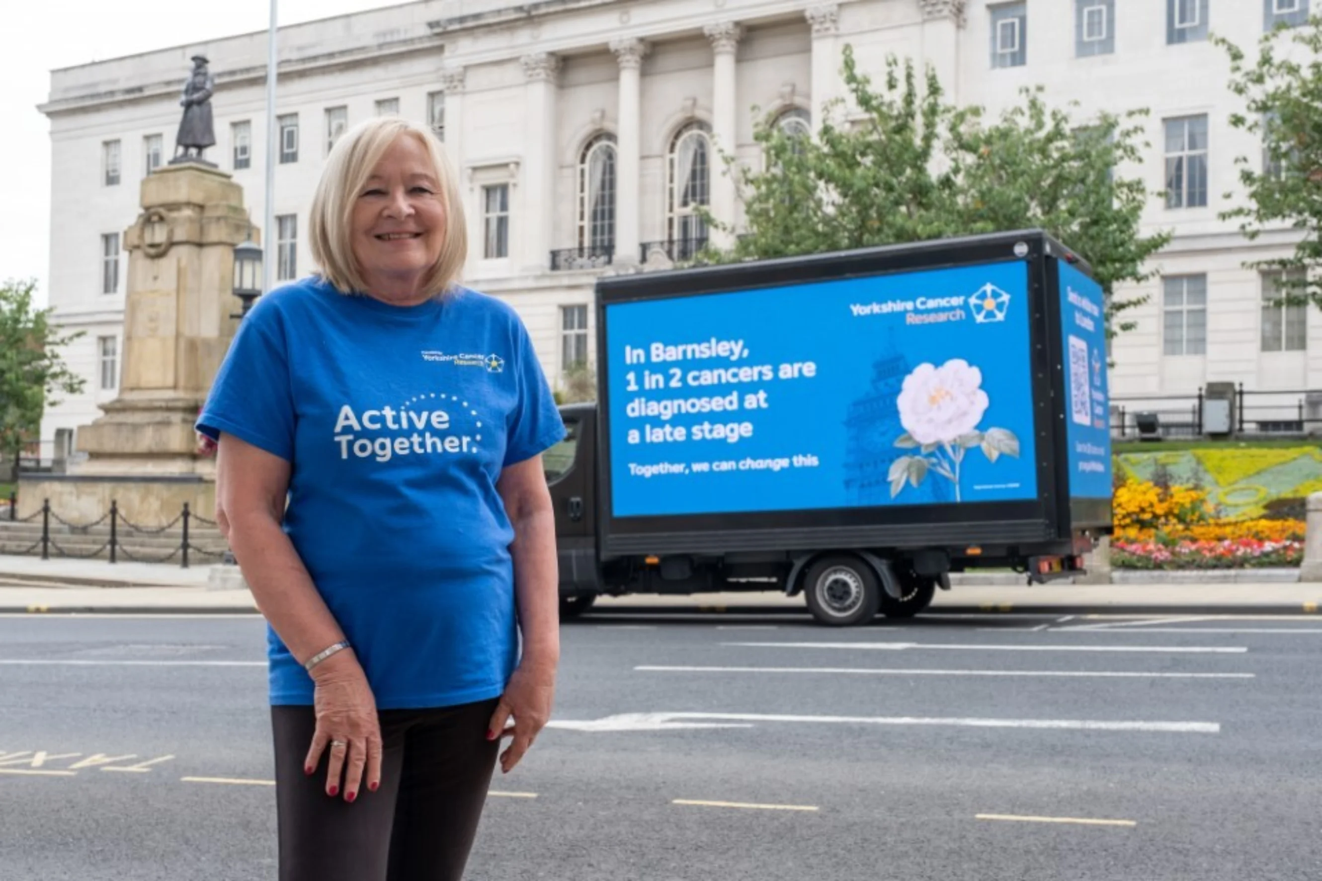 A lady wearing a blue "Active Together" t-shirt standing outside a large white building in Barnsley. behind her is a digital van that says "In Barnsley 1 in 2 cancers are diagnosed at a late stage"