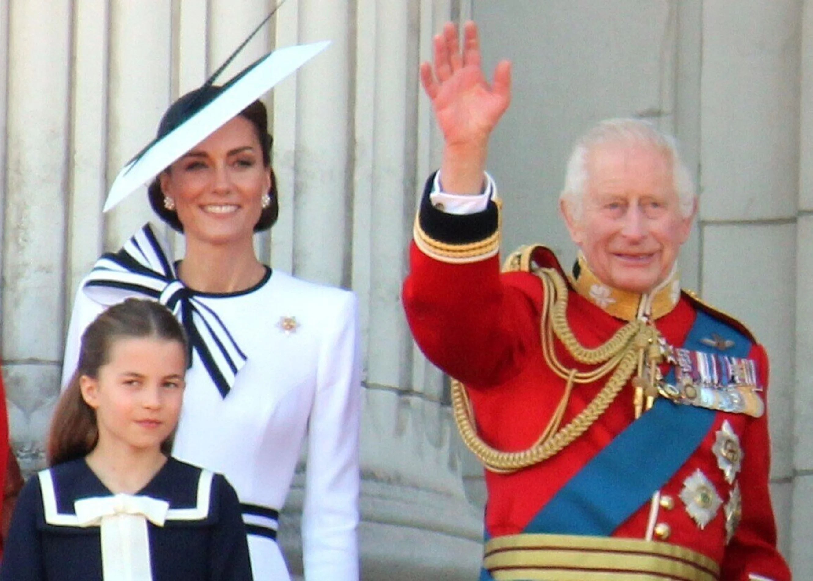 His Majesty the King and the Princess of Wales are standing waving on the balcony at Buckingham Palace.  Princess Charlotte is stood to the left of them wearing a navy and white dress.