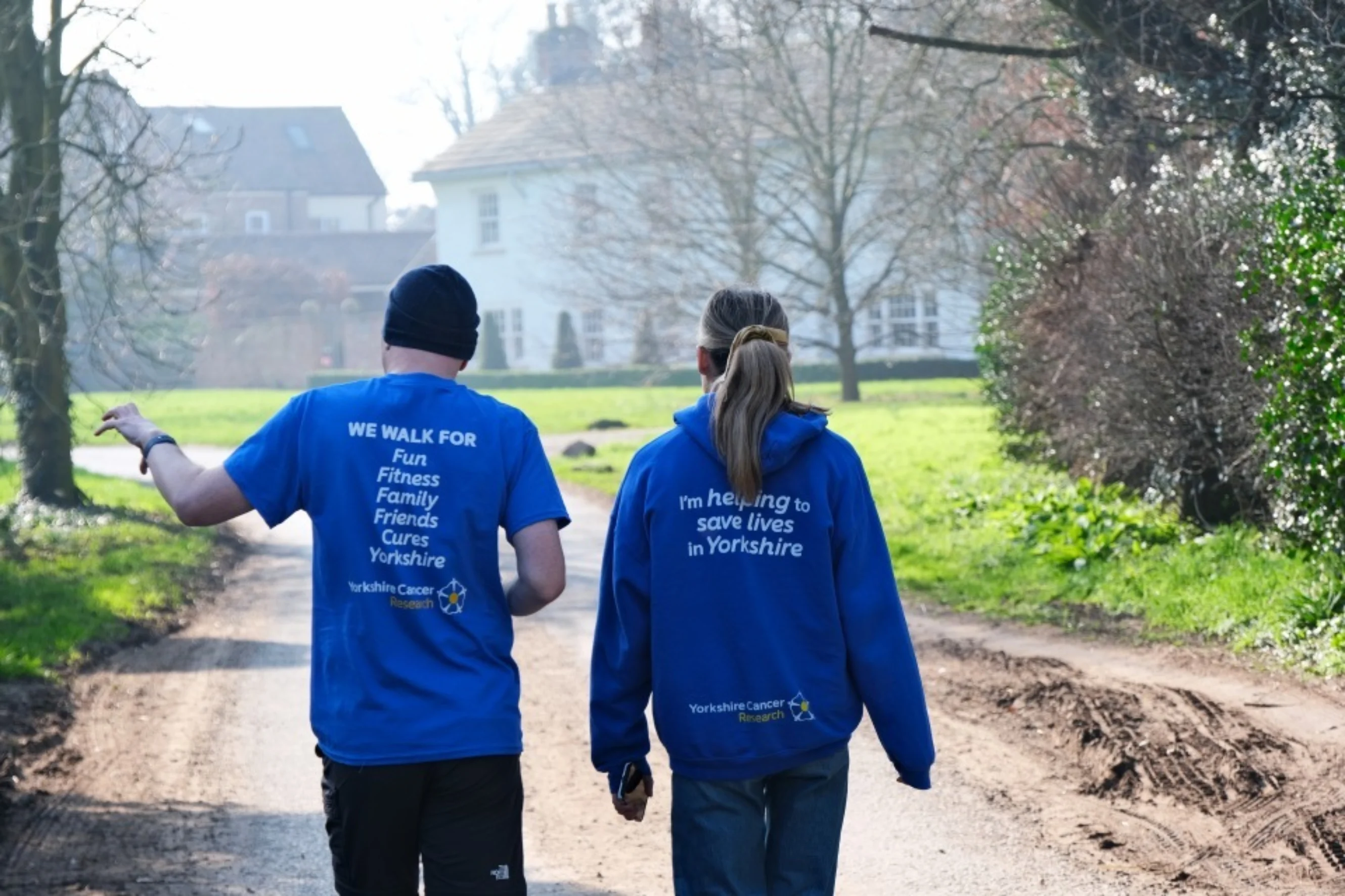Two people wearing blue branded We Walk for Yorkshire tops are walking outside on a crisp spring day. They are facing away from the camera and in front of them is a large white building and trees.