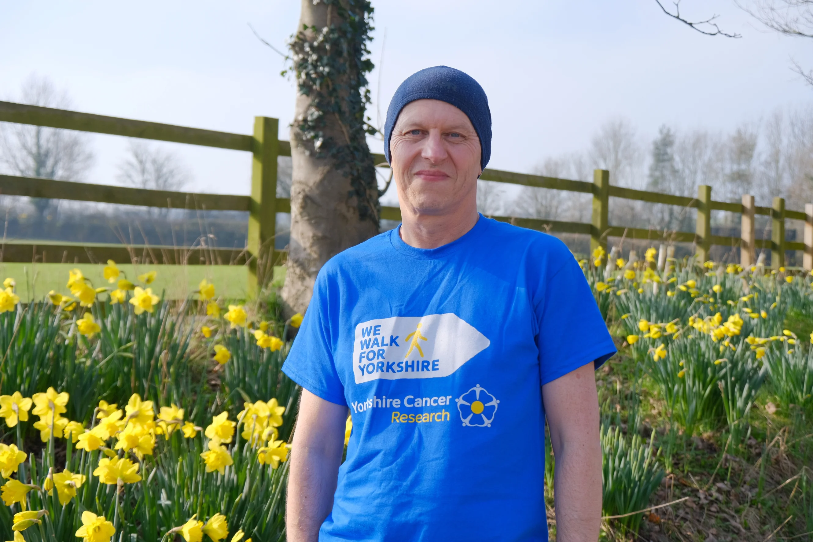 Simon is wearing a blue 'We Walk for Yorkshire' T-shirt smiling at the camera. Behind him is a grassy verge covered in daffodils.