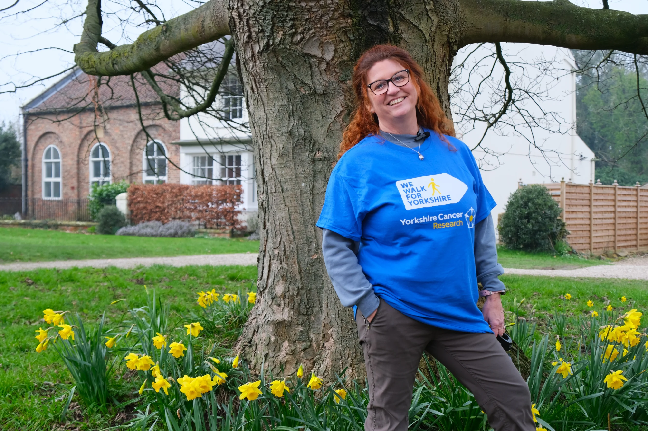 Jackie is smiling at the camera. She has red hair and is wearing black glasses. Behind her is a large tree and some daffodils.