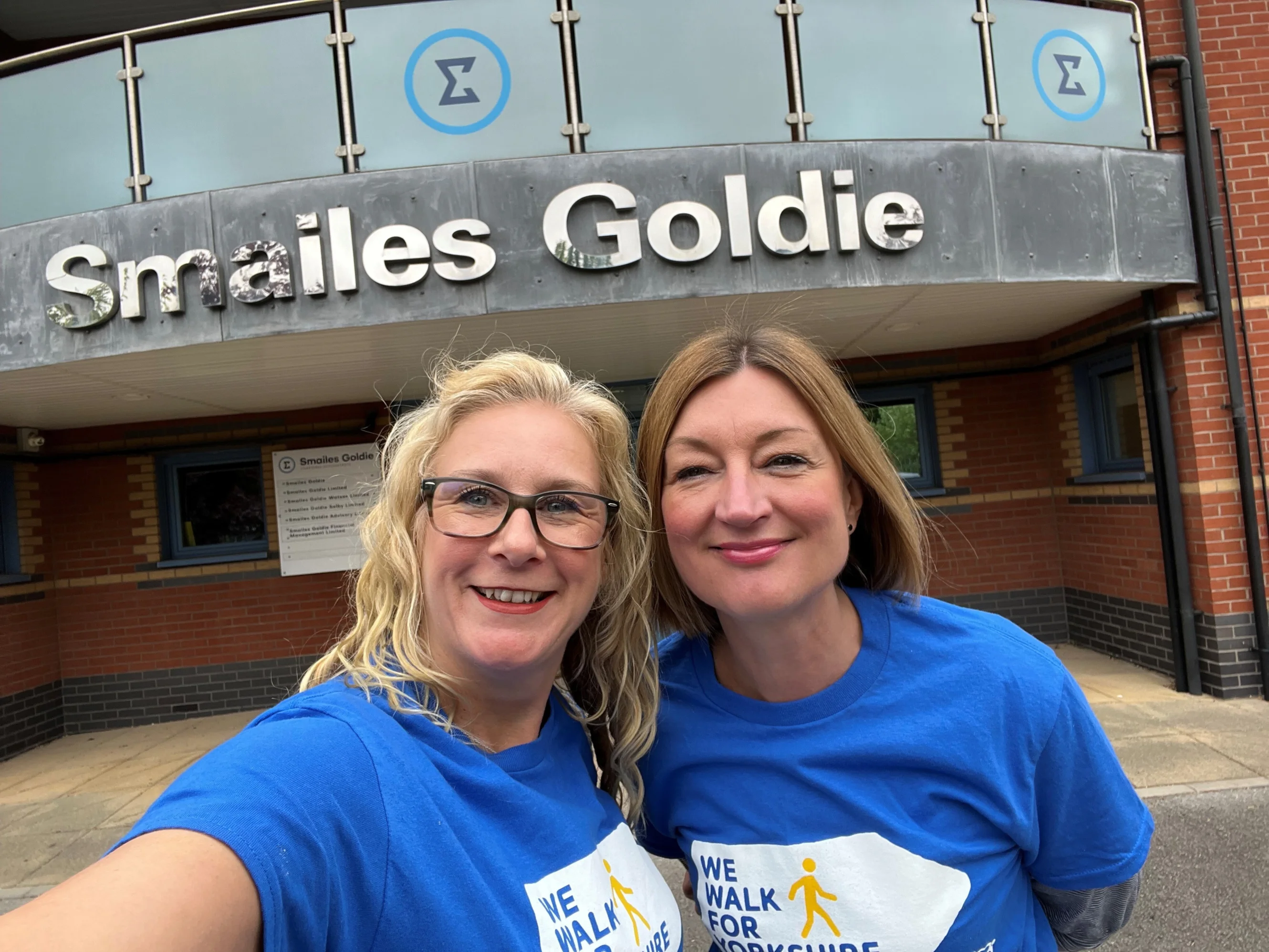 Two ladies wearing blue branded 'We Walk for Yorkshire' T-shirts in a posed selfie. Behind them is a large office building with a sign that reads "Smailes Goldie".