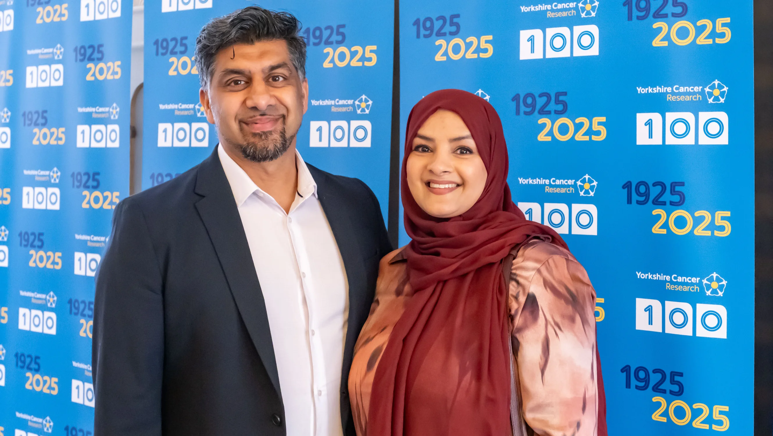 Saima is wearing a maroon hijab on the right and her husband is wearing a suit on the left. They are standing in front of Yorkshire Cancer Research 100th anniversary banners.
