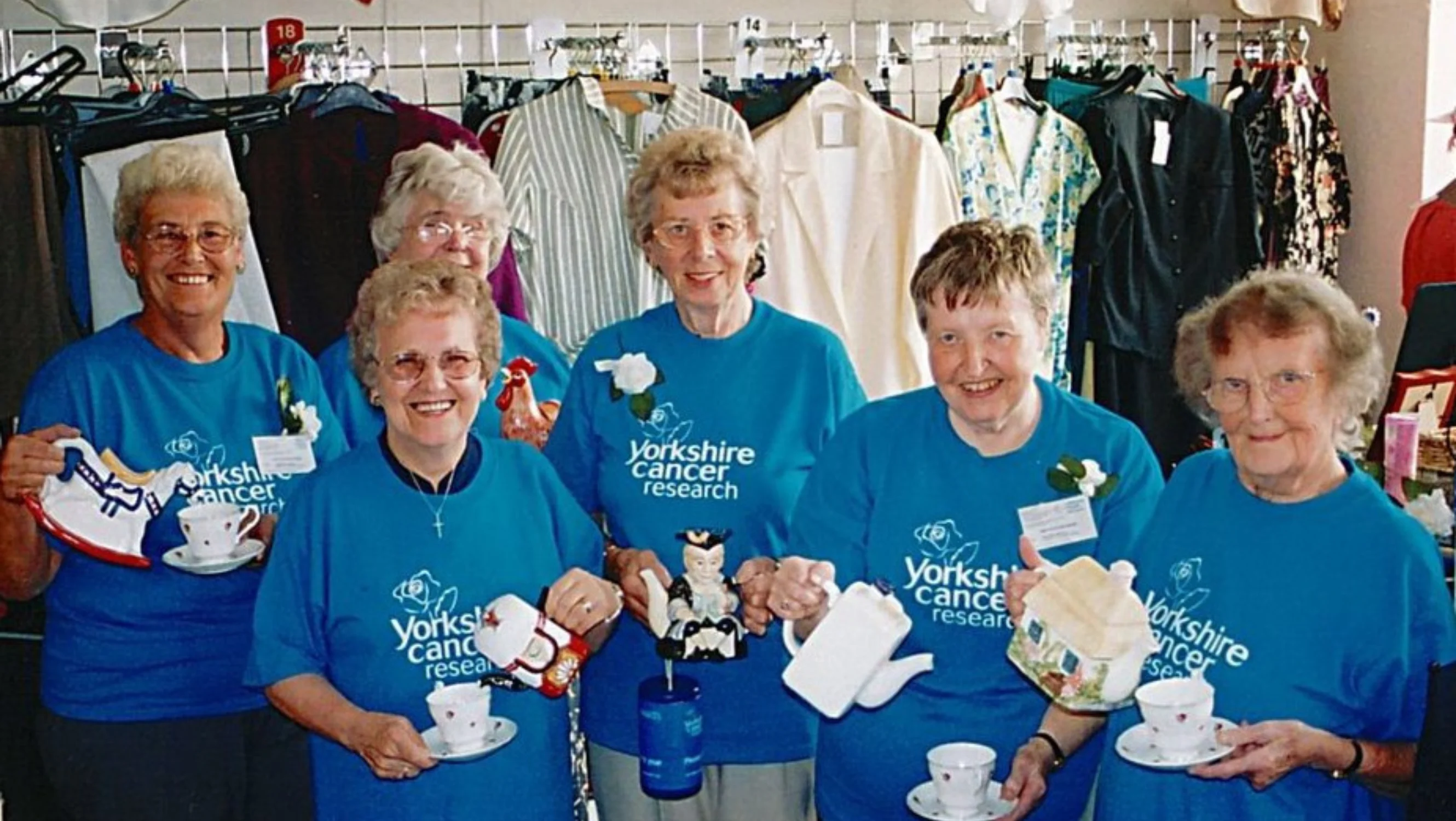 Six ladies wearing blue t-shirts holding teapots smiling at the camera. They are stood in a retail setting.