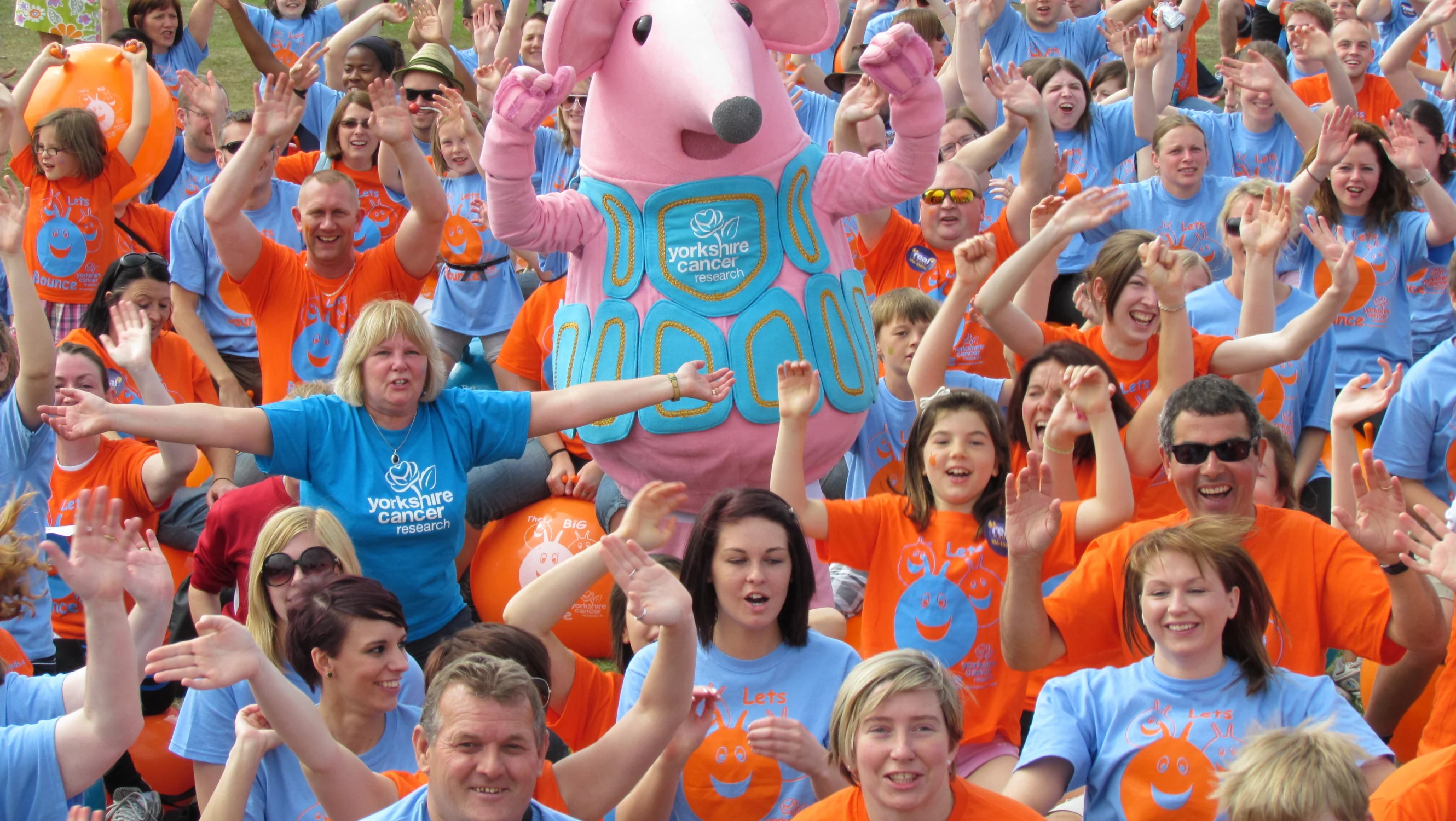 A crowd of people all waving and smiling while sitting on space hoppers at a fundraising event.