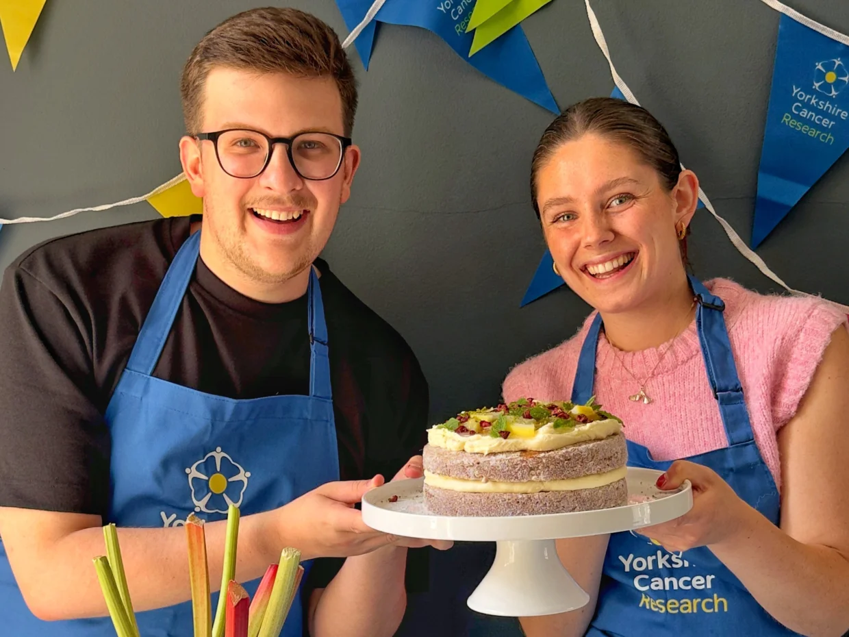 Two people holding a rhubarb doughnut cake. They are wearing blue aprons and behind them is bright charity-branded bunting.