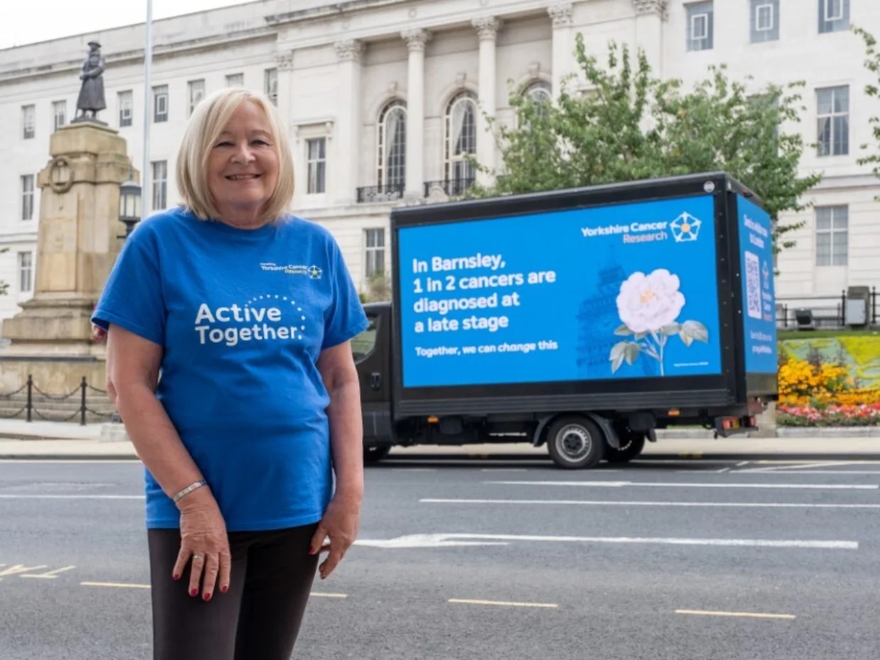 A lady wearing a blue "Active Together" t-shirt standing outside a large white building in Barnsley. behind her is a digital van that says "In Barnsley 1 in 2 cancers are diagnosed at a late stage"