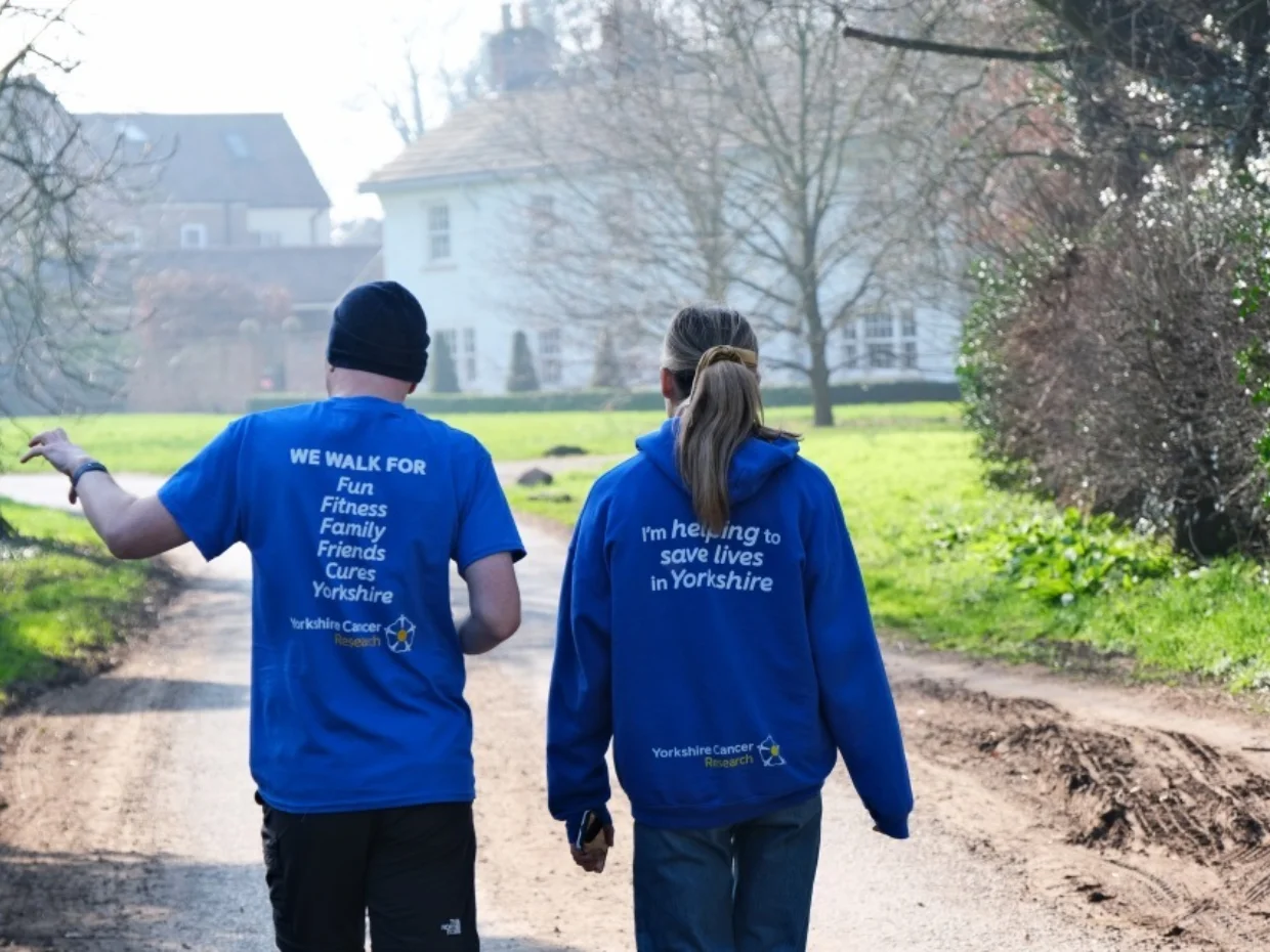 Two people wearing blue branded We Walk for Yorkshire tops are walking outside on a crisp spring day. They are facing away from the camera and in front of them is a large white building and trees.