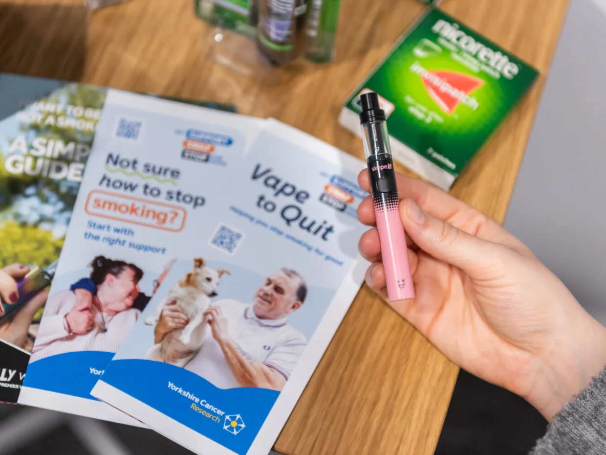 A hand holding a pink vape. On the table in the background are leaflets about stop smoking support