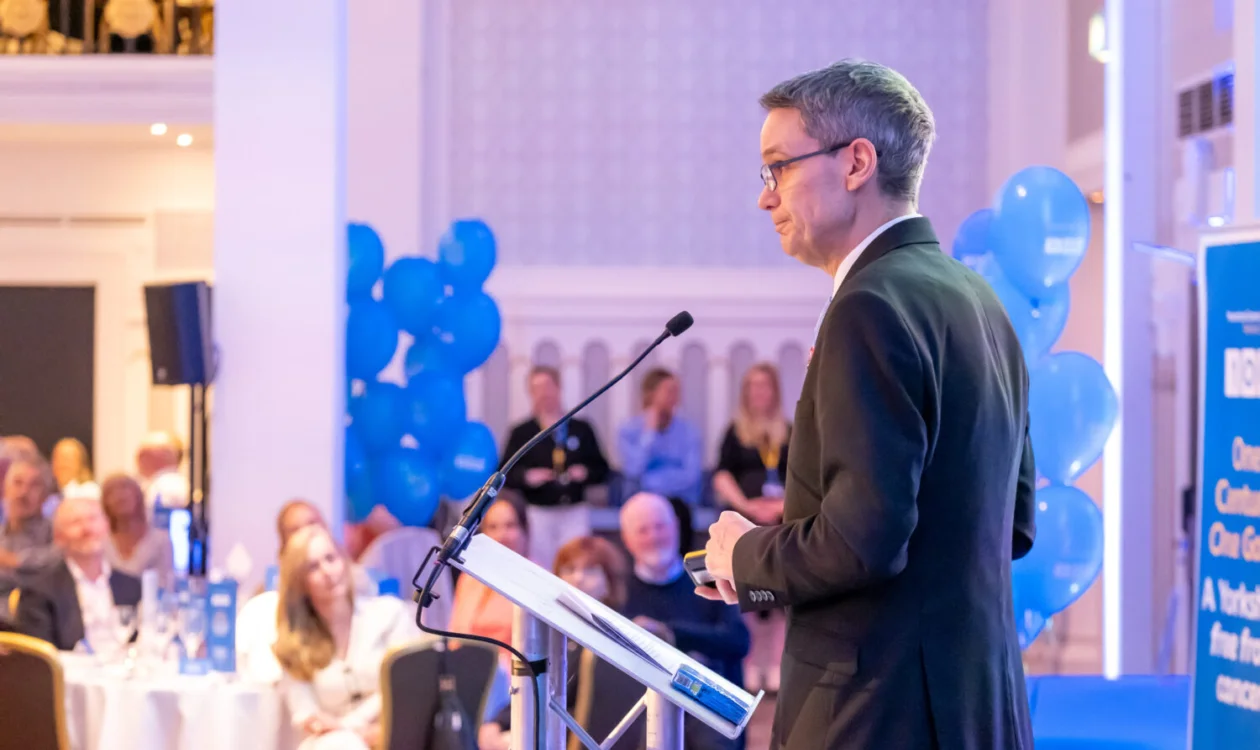A man stands in front of a lectern and microphone speaking to a room of people