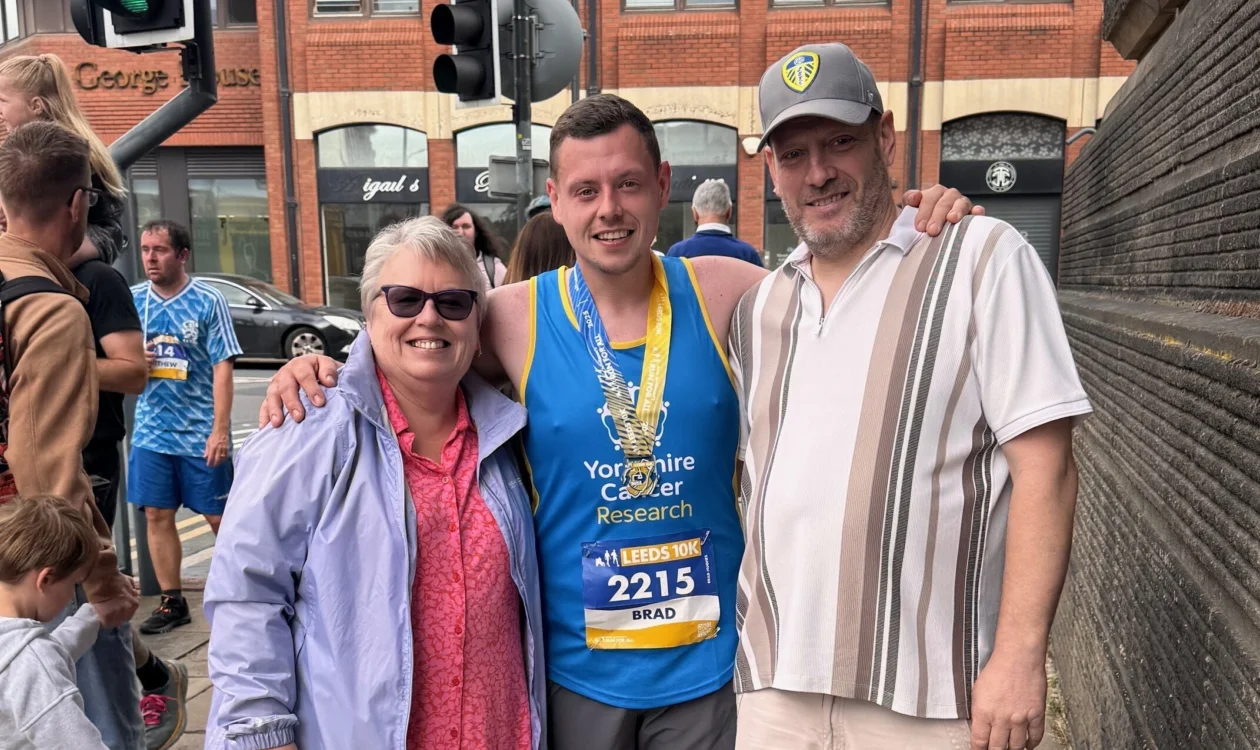 Brad is stood in the street in his running gear with his medal. His standing with two other people. They are all smiling at the camera.