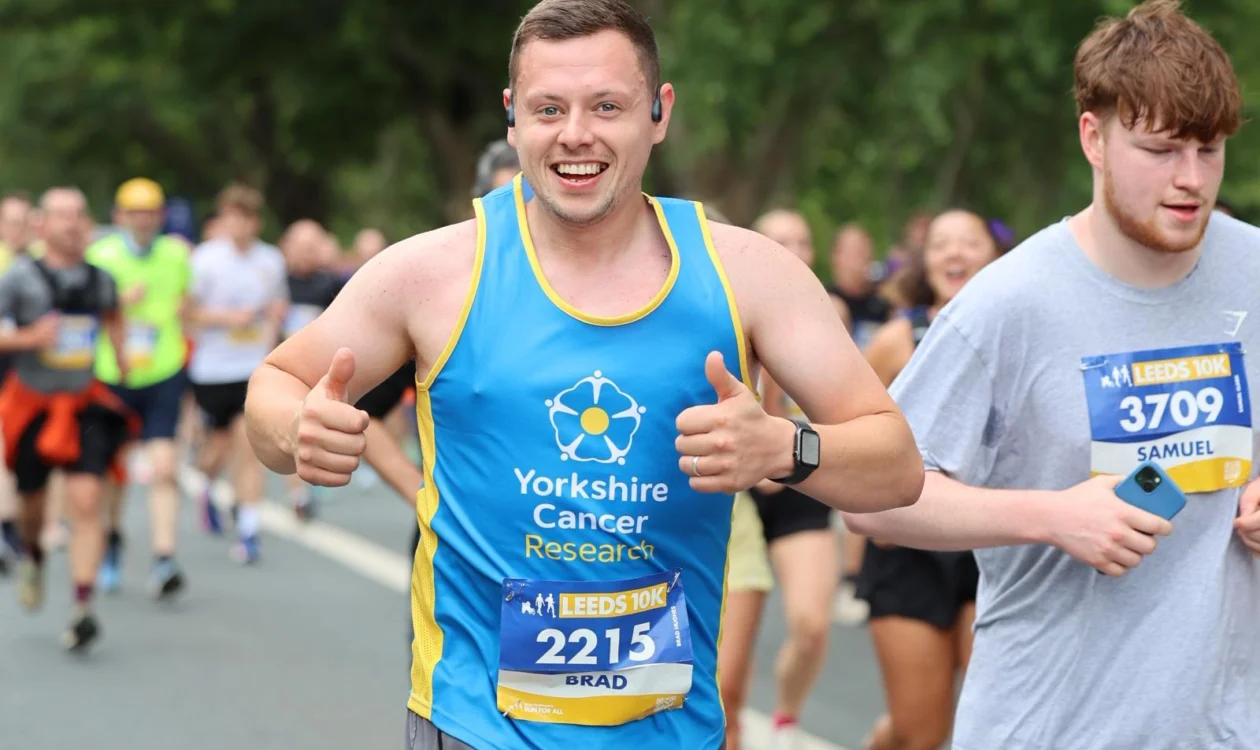 Brad is running in the Leeds 10k and smiling happily at the camera. He is wearing a blue Yorkshire Cancer Research vest.