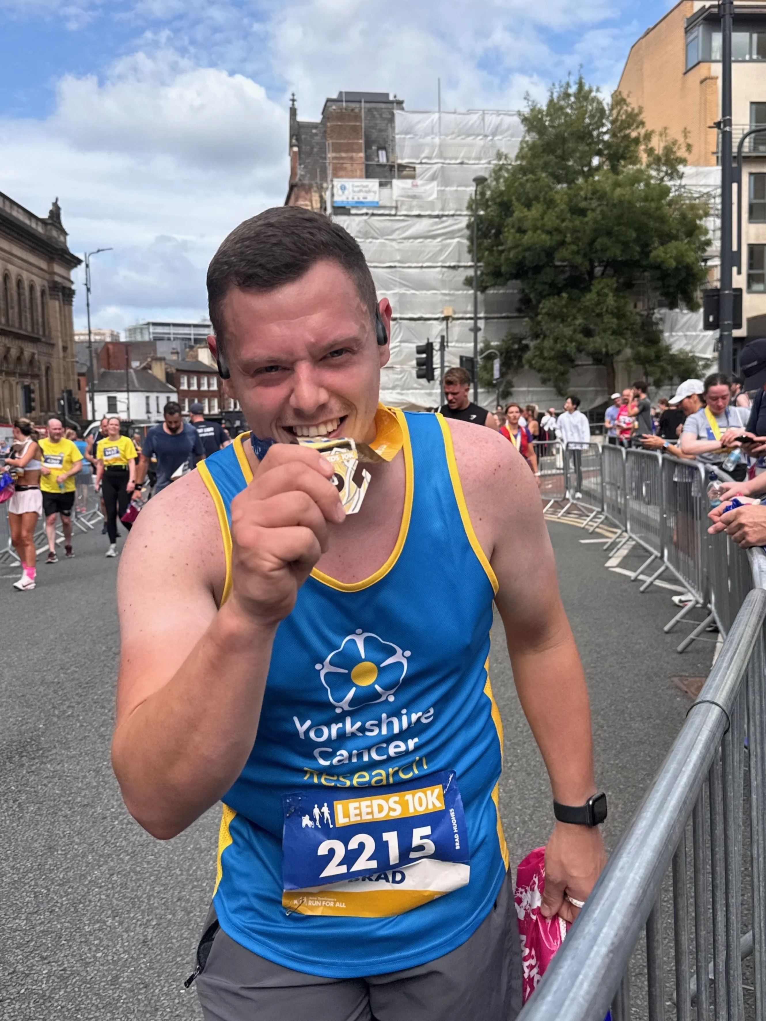Brad is stood to the side-lines of the finish line. He is biting his medal.
