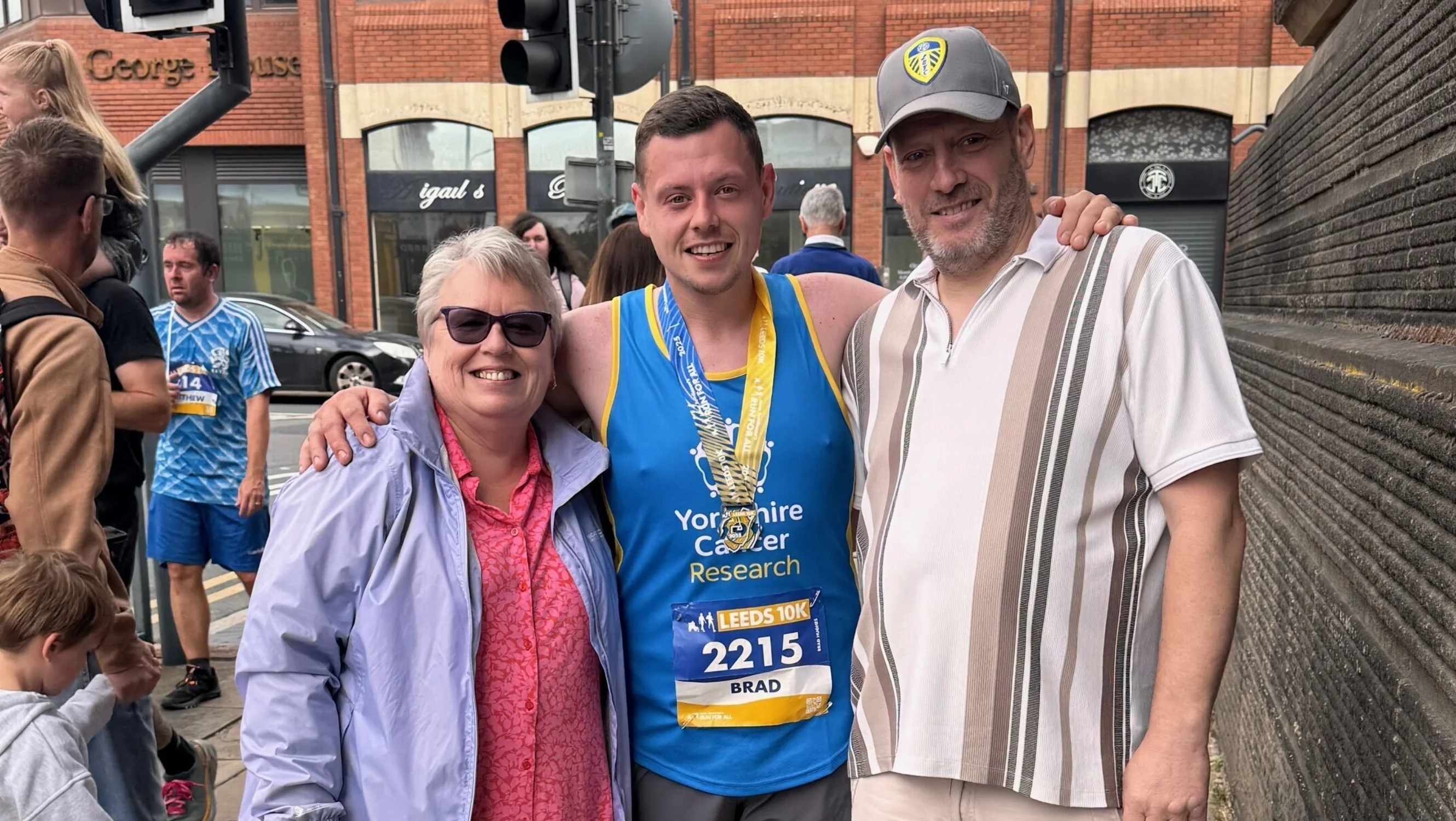 Brad is stood in the street in his running gear with his medal. His standing with two other people. They are all smiling at the camera.
