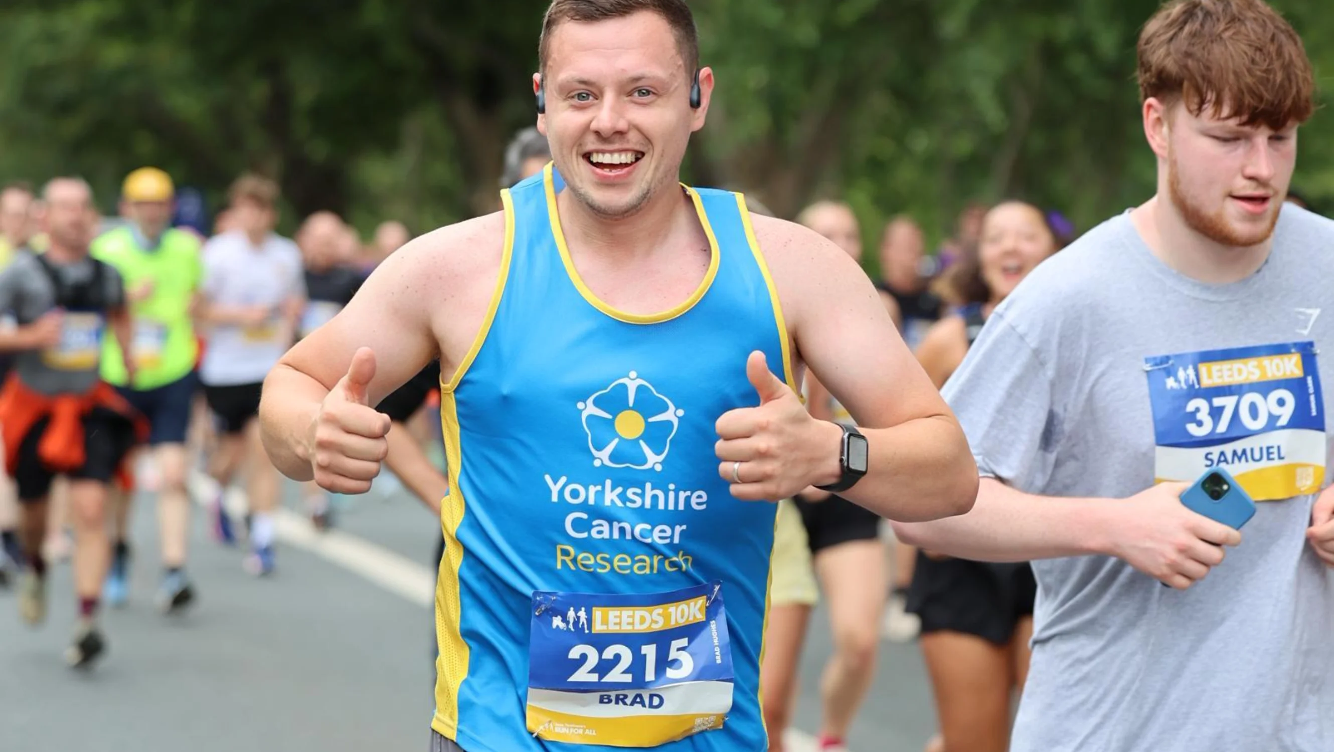Brad is running in the Leeds 10k and smiling happily at the camera. He is wearing a blue Yorkshire Cancer Research vest.