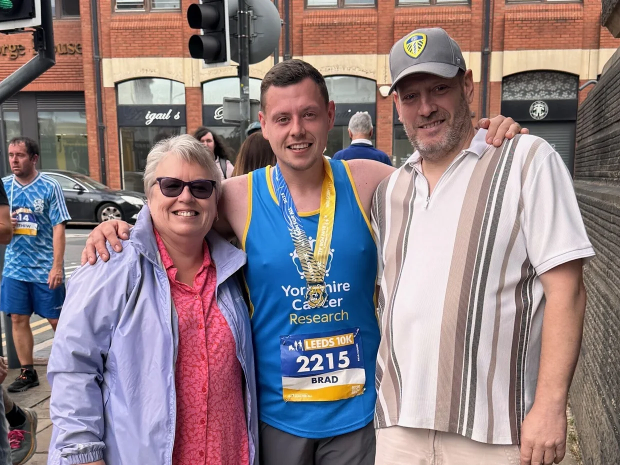 Brad is stood in the street in his running gear with his medal. His standing with two other people. They are all smiling at the camera.