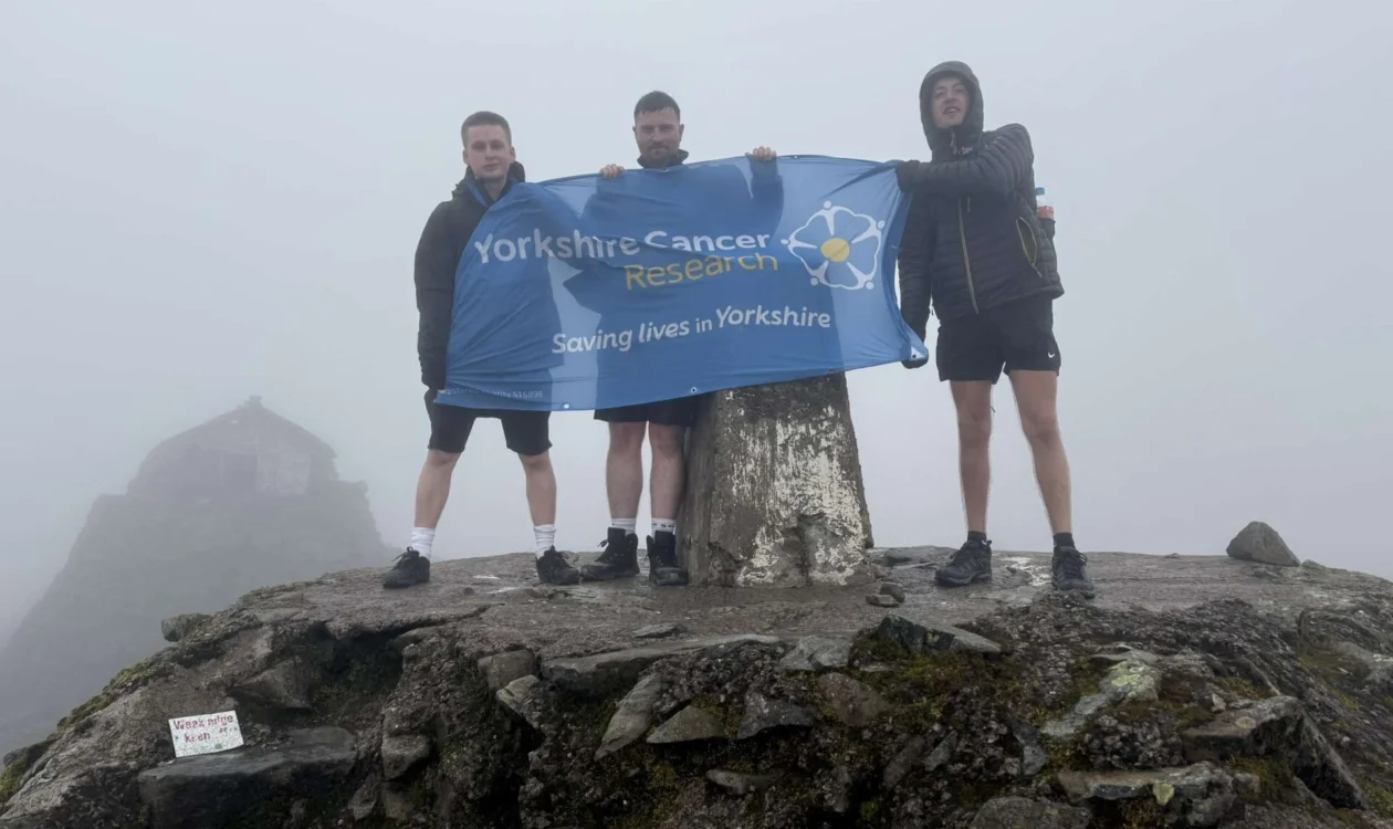 Three people stood at the peak of Ben Nevis holding  Yorkshire Cancer Research banner.