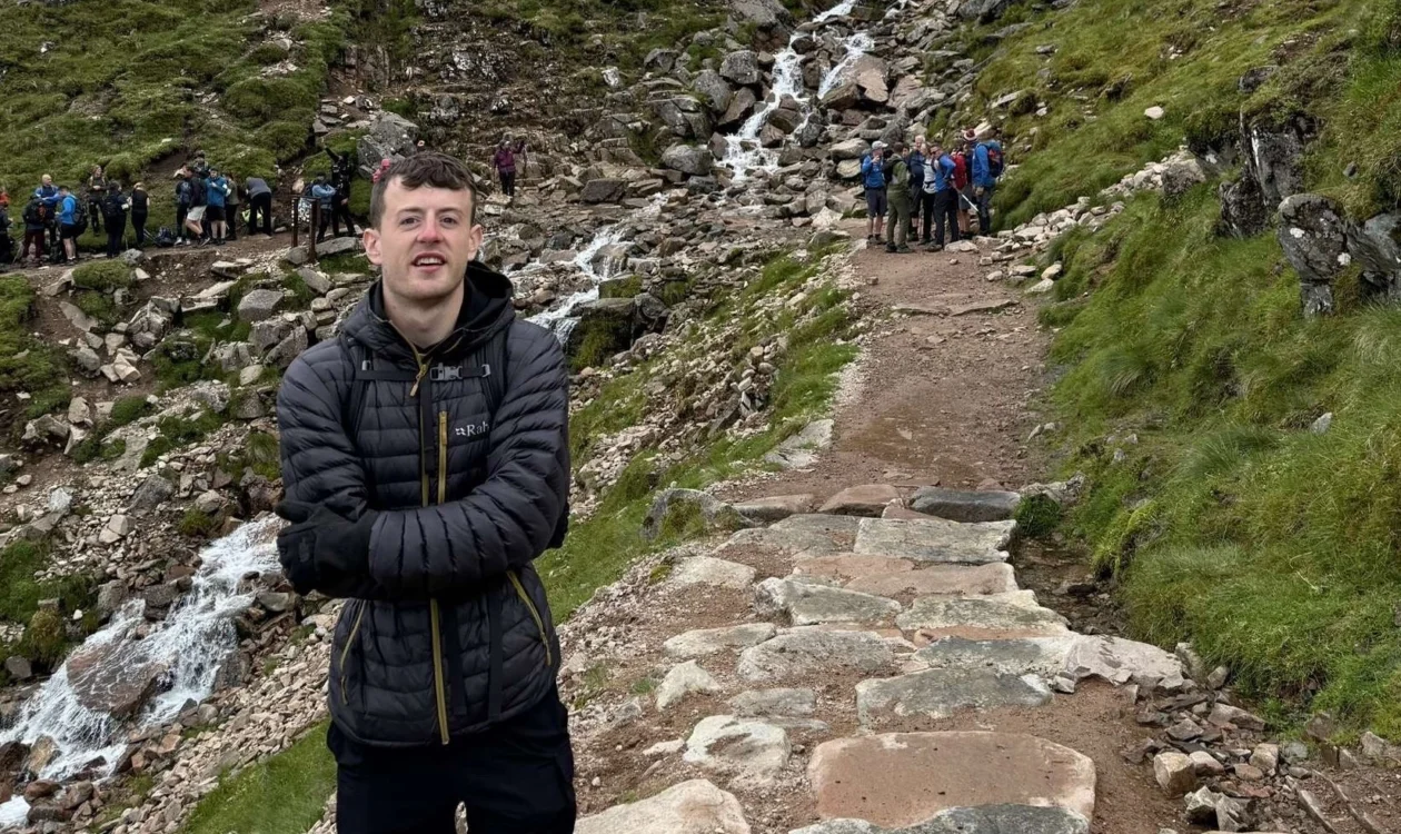Lewis is stood on a climbing trail along Ben Nevis wearing a black jacket. Behind him is a line of people and green hills.