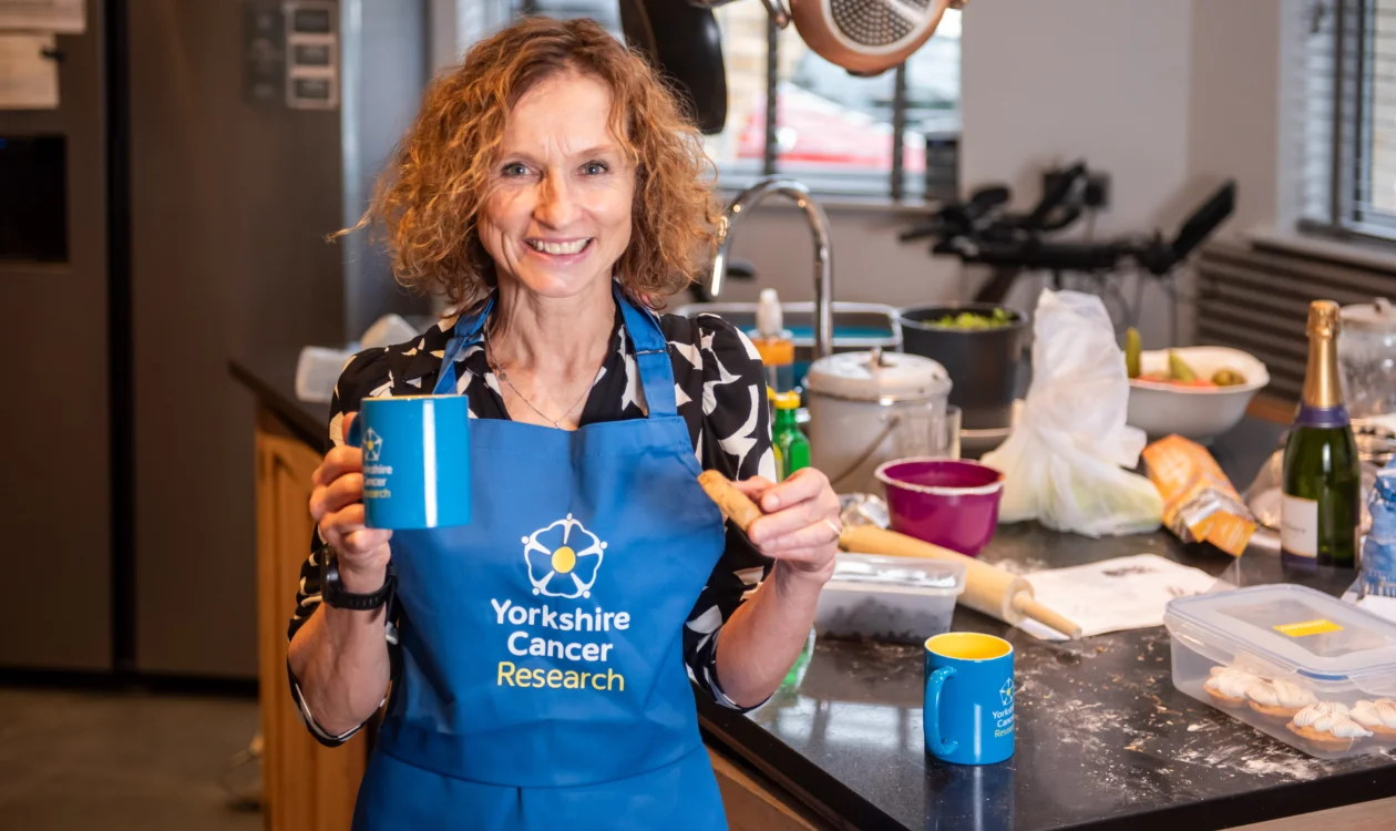 Jackie is stood in a kitchen wearing a blue Yorkshire Cancer Research apron holding a mug and a biscuit.