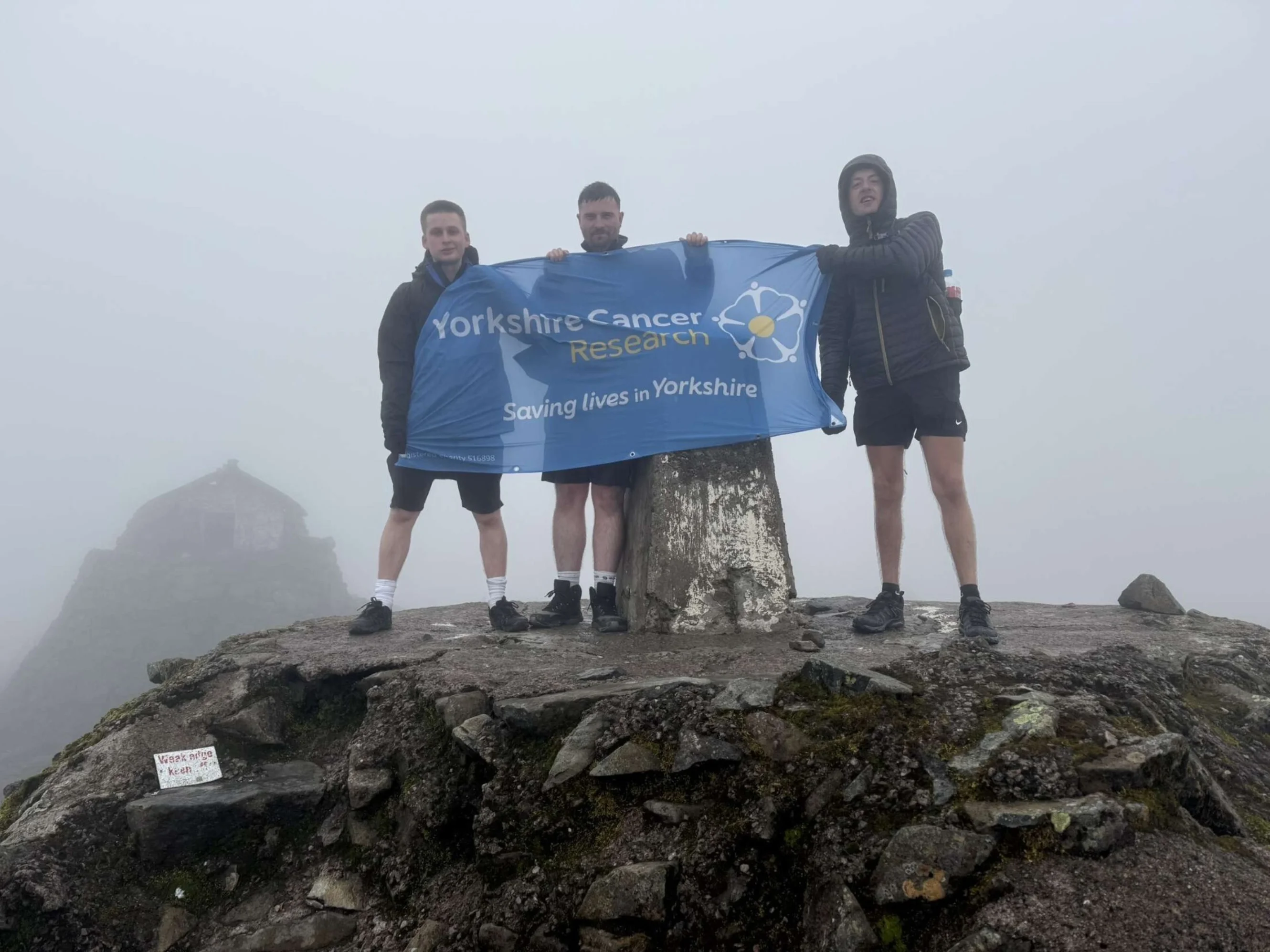 Three people stood at the peak of Ben Nevis holding Yorkshire Cancer Research banner.