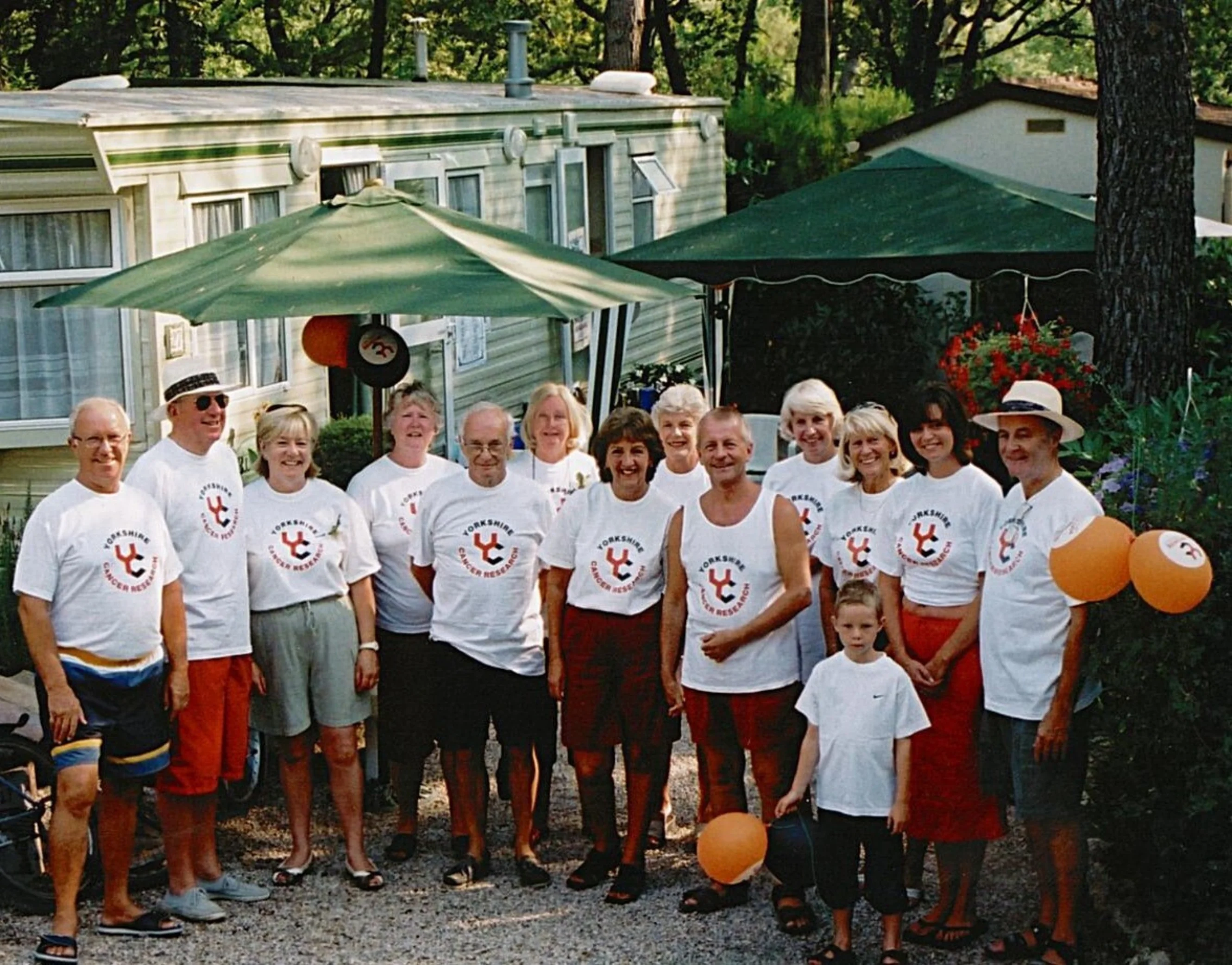 14 people are stood outside a static caravan wearing white t-shirts with the charity's old logo on. Some are holding balloons.