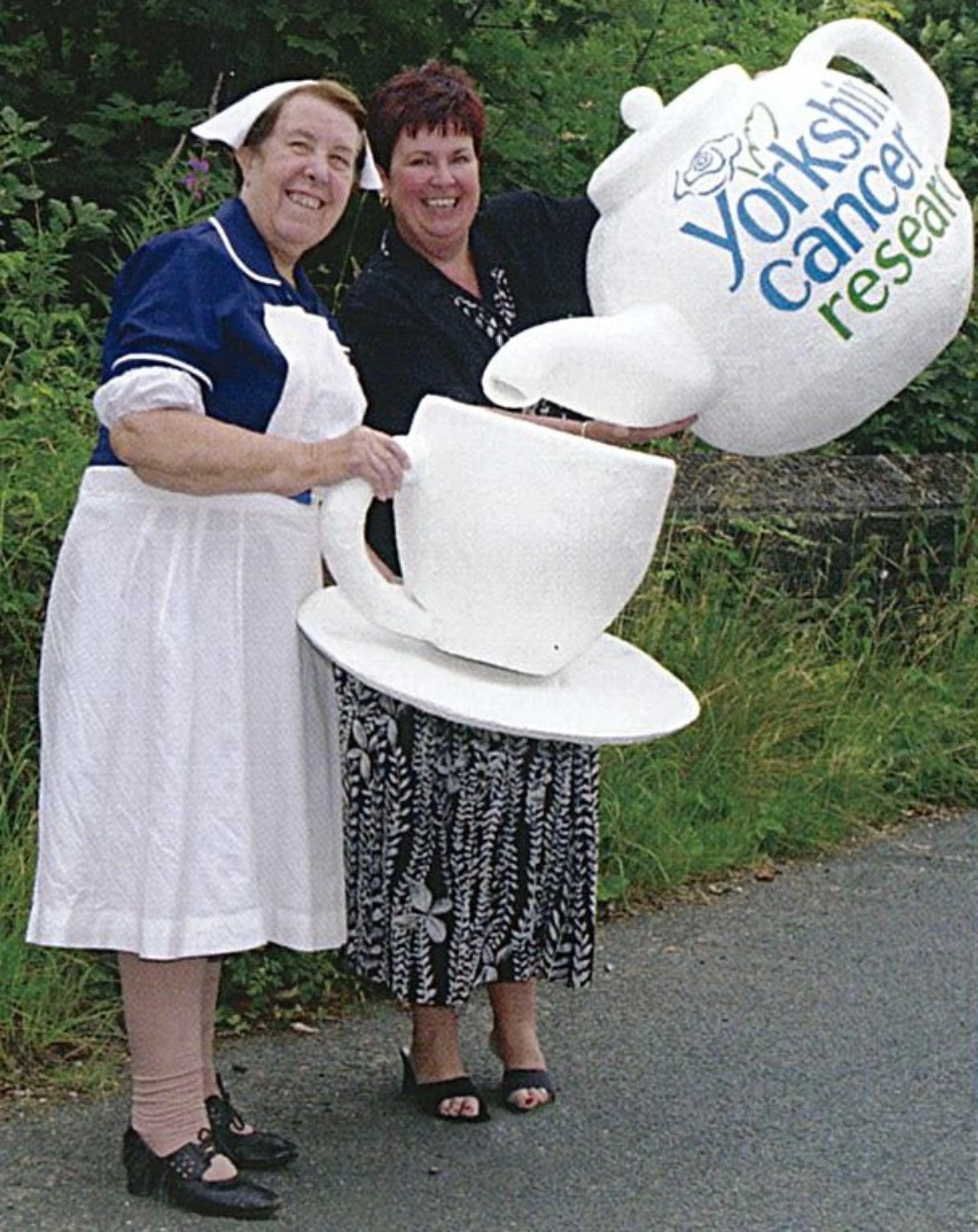 Kathy staff helped the Yorkshire Cancer Research Huddersfield LVG support Yorkshires Biggest Teabreak. She is holding a giant mug while someone else is pouring a giant teapot with the charity's logo on it.