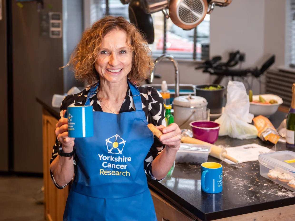 Jackie is stood in a kitchen wearing a blue Yorkshire Cancer Research apron holding a mug and a biscuit.