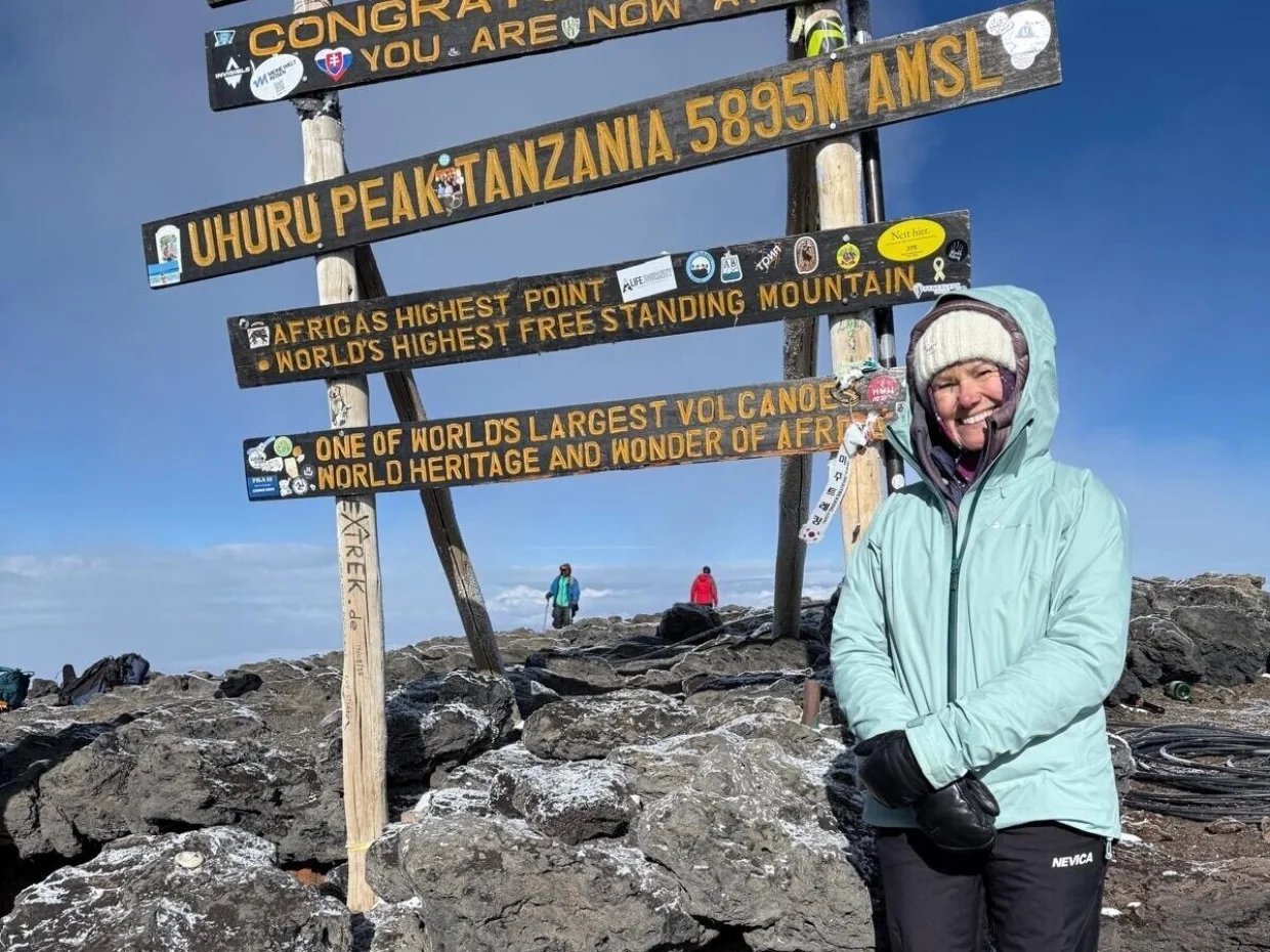 Imogen is wearing a blue coat and white hat. She is standing at the top of Mt Kilimanjaro next to a large sign. In the background you can see clouds and a blue sky.