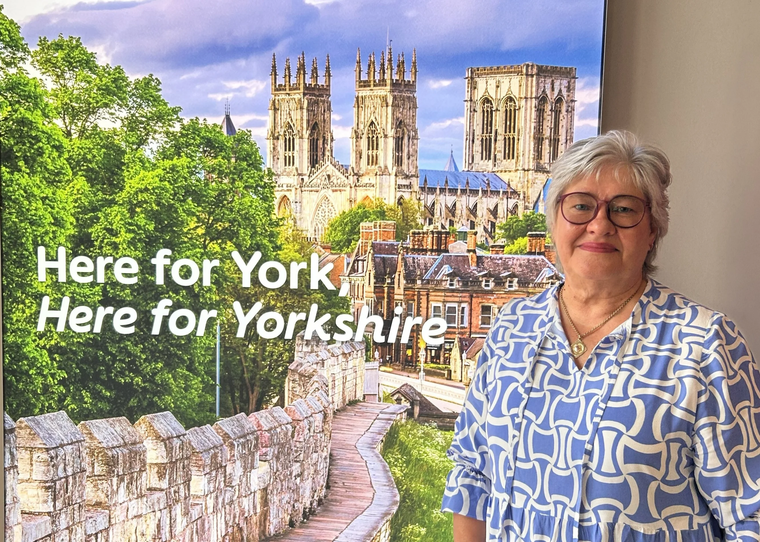 Christine wears a blue patterned dress and stands next to a large lit up photo of York Minster and the city walls. The text "Here for York, Here for Yorkshire" is also visible.
