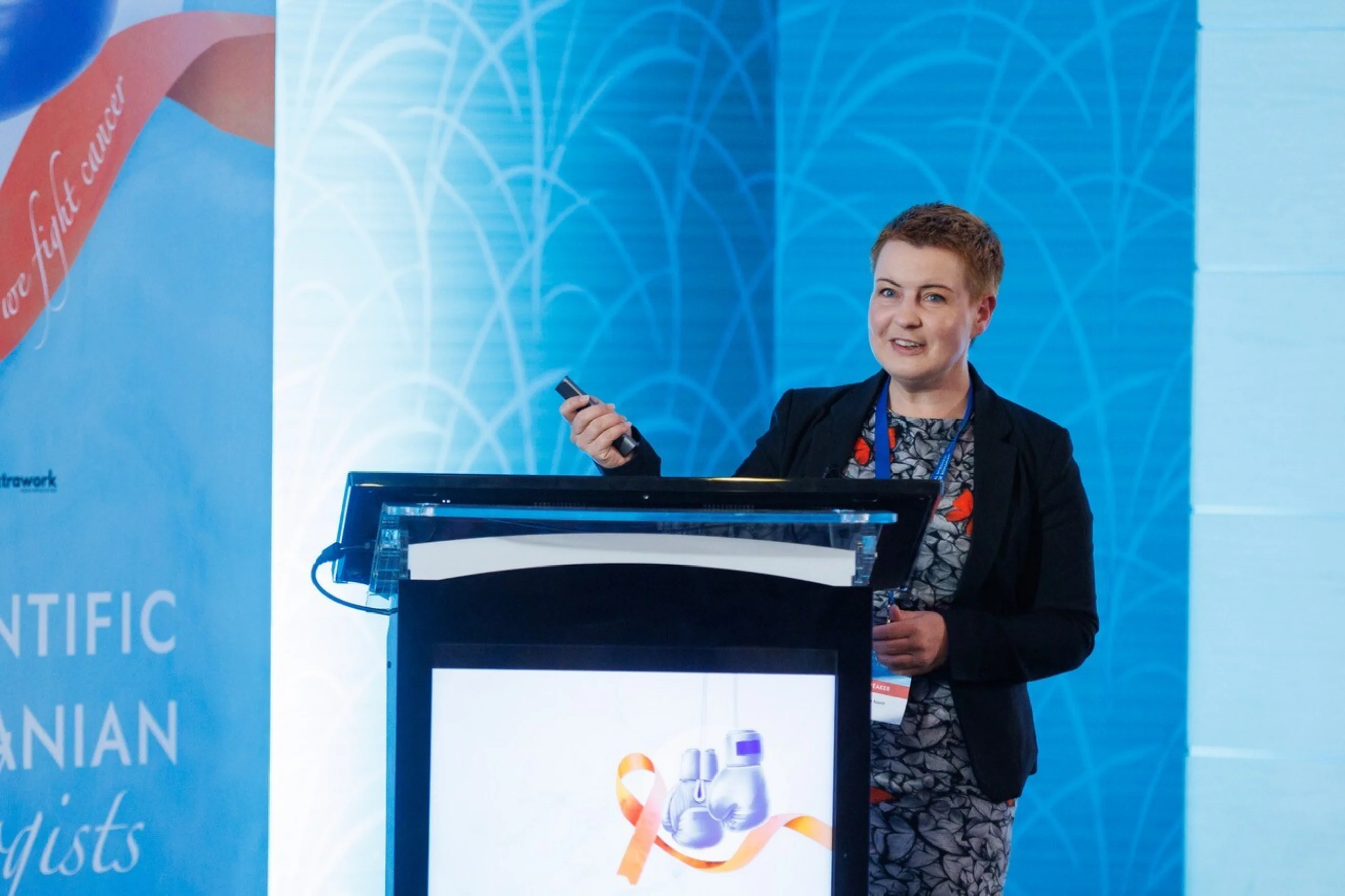 Ane stands behind a podium at a conference, smiling and holding a microphone.