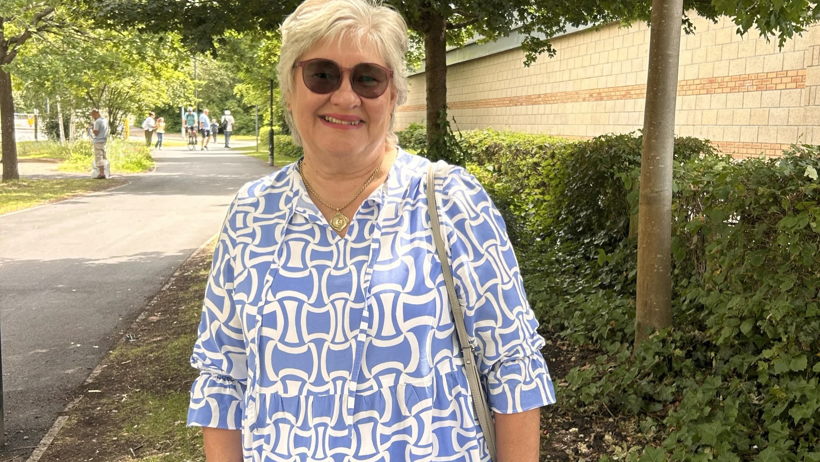 Christine wears a patterned blue dress and sunglasses while standing outdoors on a sunny day. A footpath and trees are visible nearby.