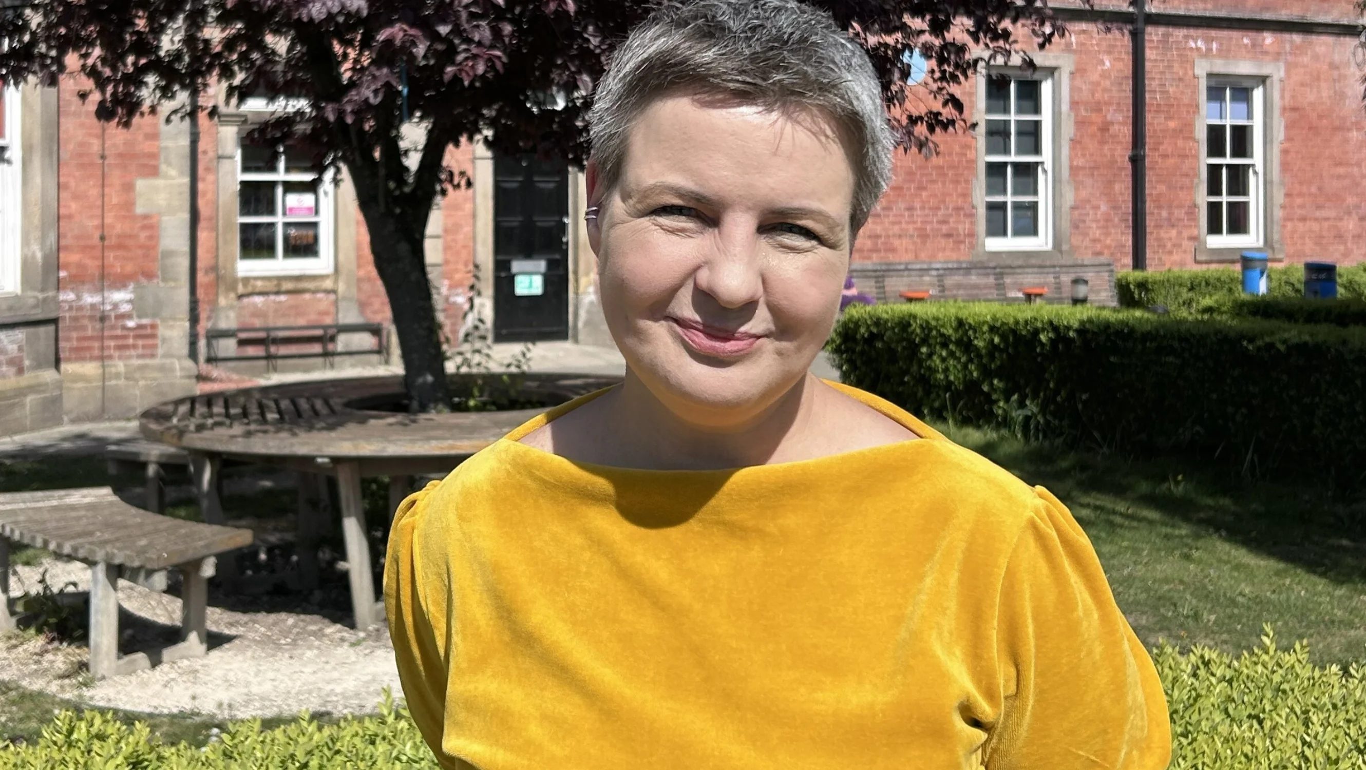 Ane wears a yellow shirt and stands outdoors on a sunny day. A building, tree and bench are visible in the background.