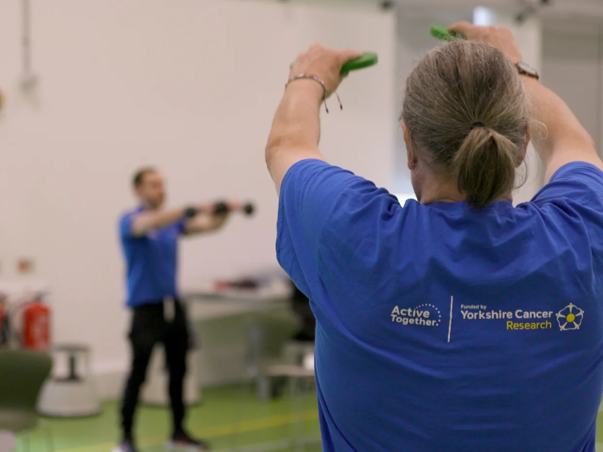 A man in a blue branded Active Together shirt is lifting weights with his back to the camera. In the background you can see a fitness instructor.
