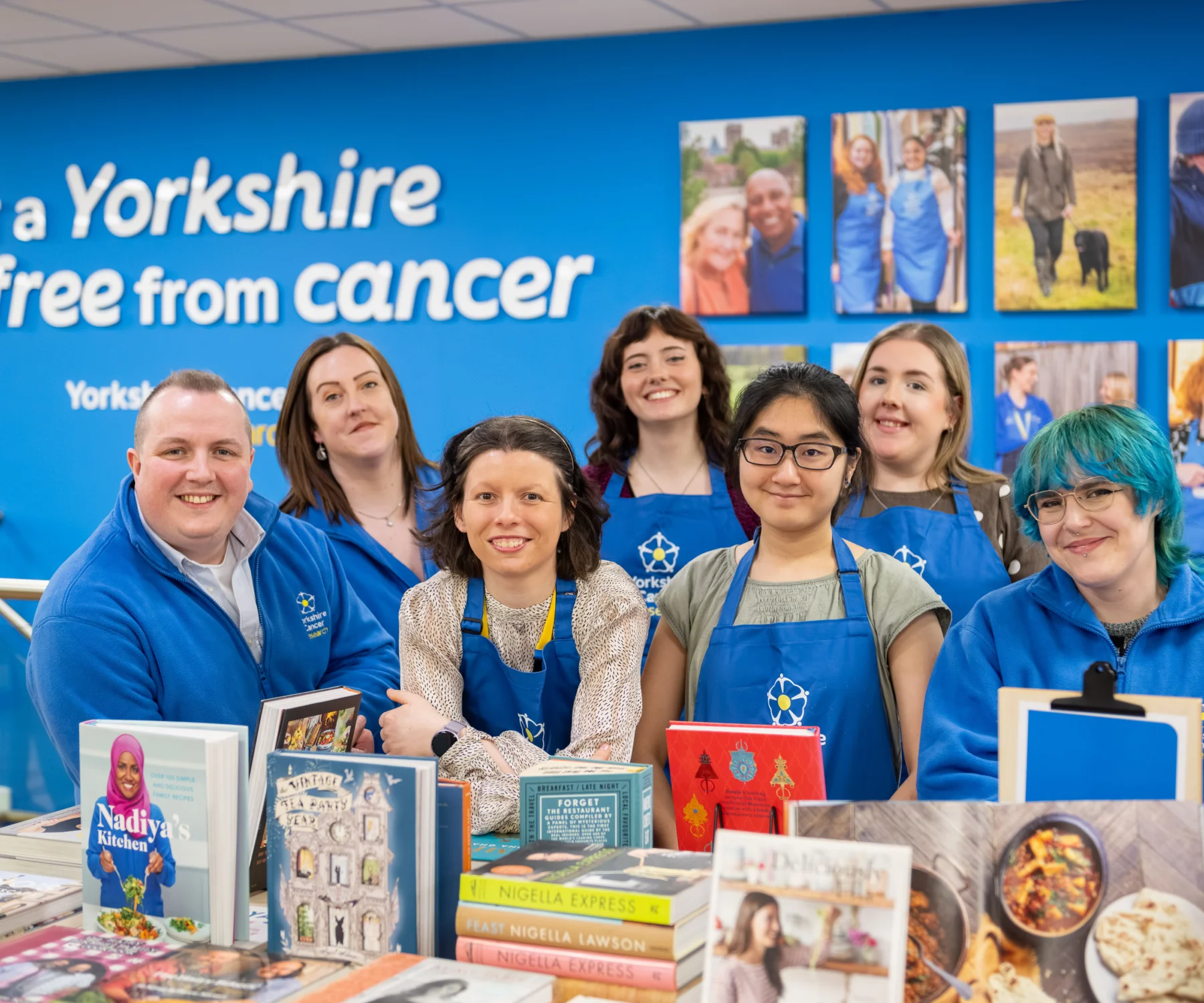 A team of seven retail staff wearing blue Yorkshire Cancer Research aprons and hoodies are stood around a table of books in the shop. Behind them is a blue wall with photos of various  volunteers and fundraisers and a sign that reads "For a Yorkshire free from cancer"