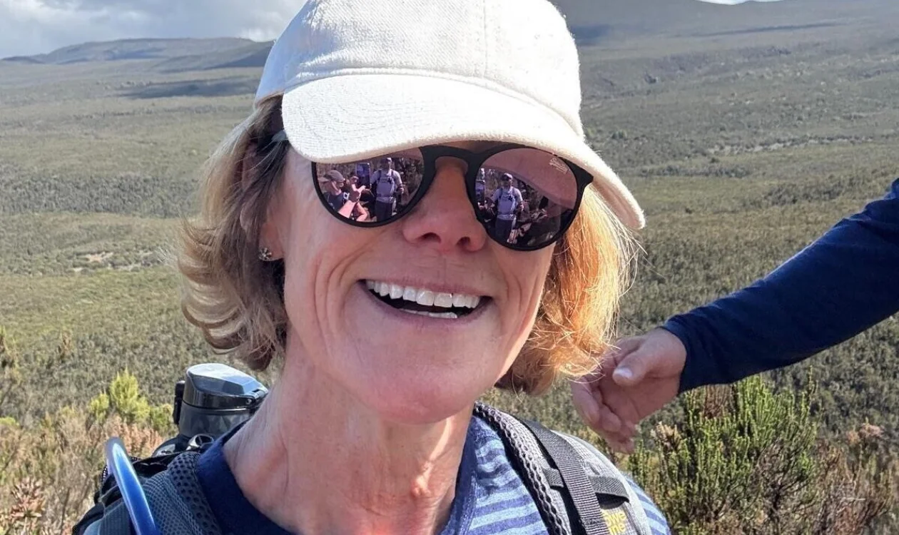 Imogen wearing sunglasses and a beige cap taking a selfie while climbing Mt Kilimanjaro. In the background are large hills.