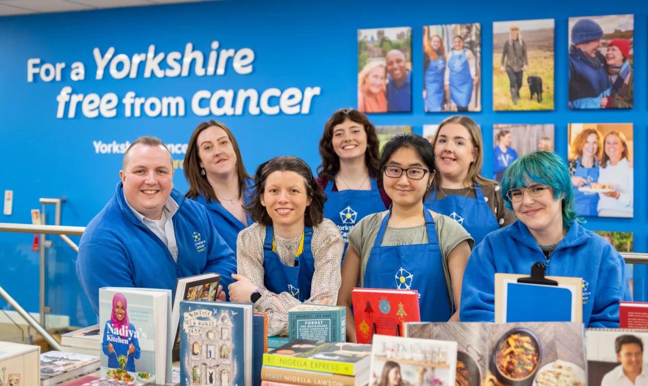 A team of seven retail staff wearing blue Yorkshire Cancer Research aprons and hoodies are stood around a table of books in the shop. Behind them is a blue wall with photos of various  volunteers and fundraisers and a sign that reads "For a Yorkshire free from cancer"