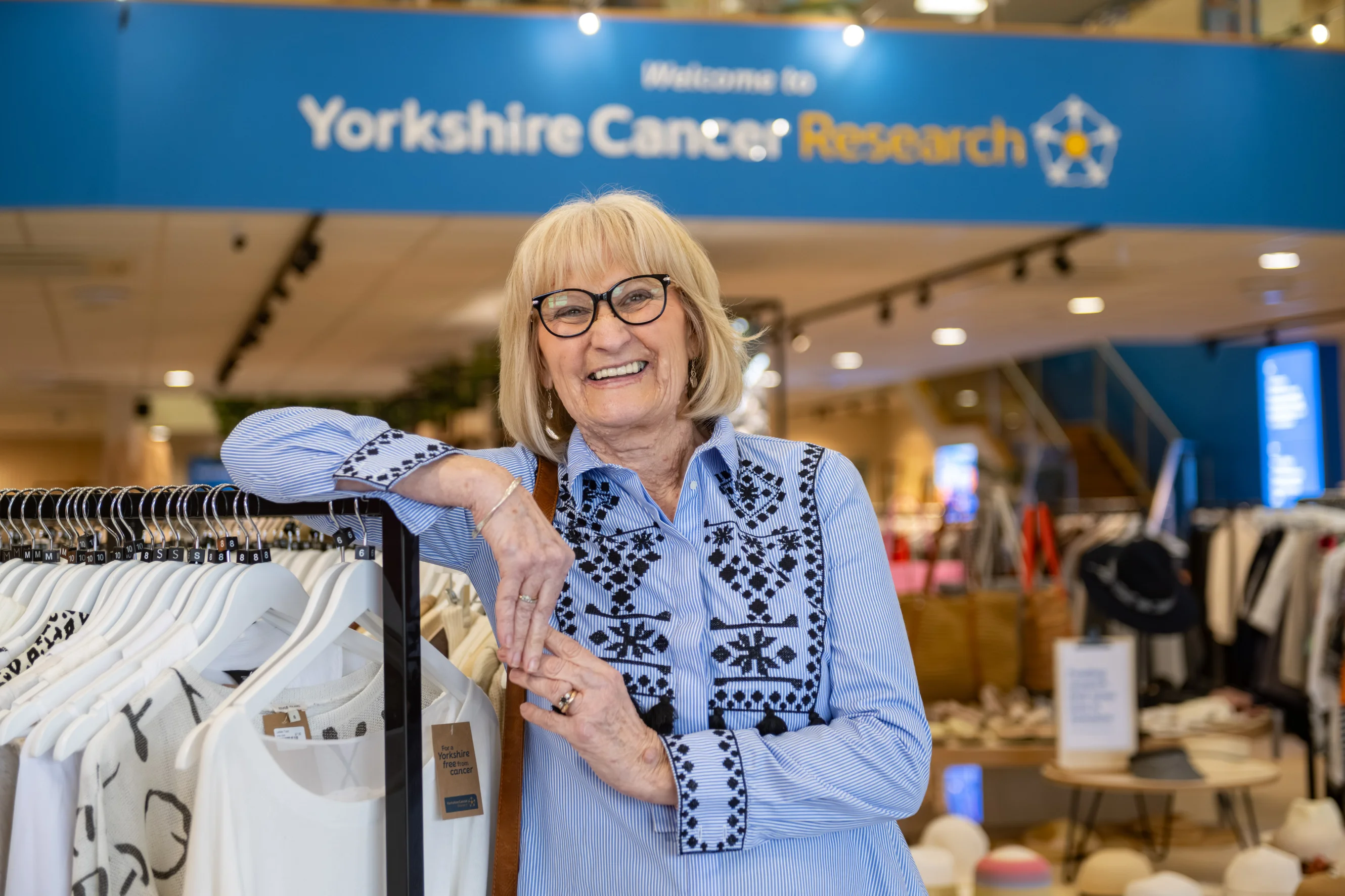 Chris is wearing a blue embroidered blouse and is leaning on a clothing rail in the shop. She is smiling at the camera and wearing black glasses. Behind her is a sign that with the charity's logo on it.